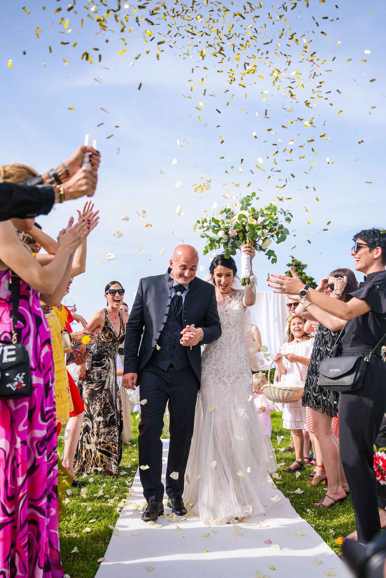 Newlyweds walk down aisle as guests shower them with golden confetti at outdoor wedding celebration.