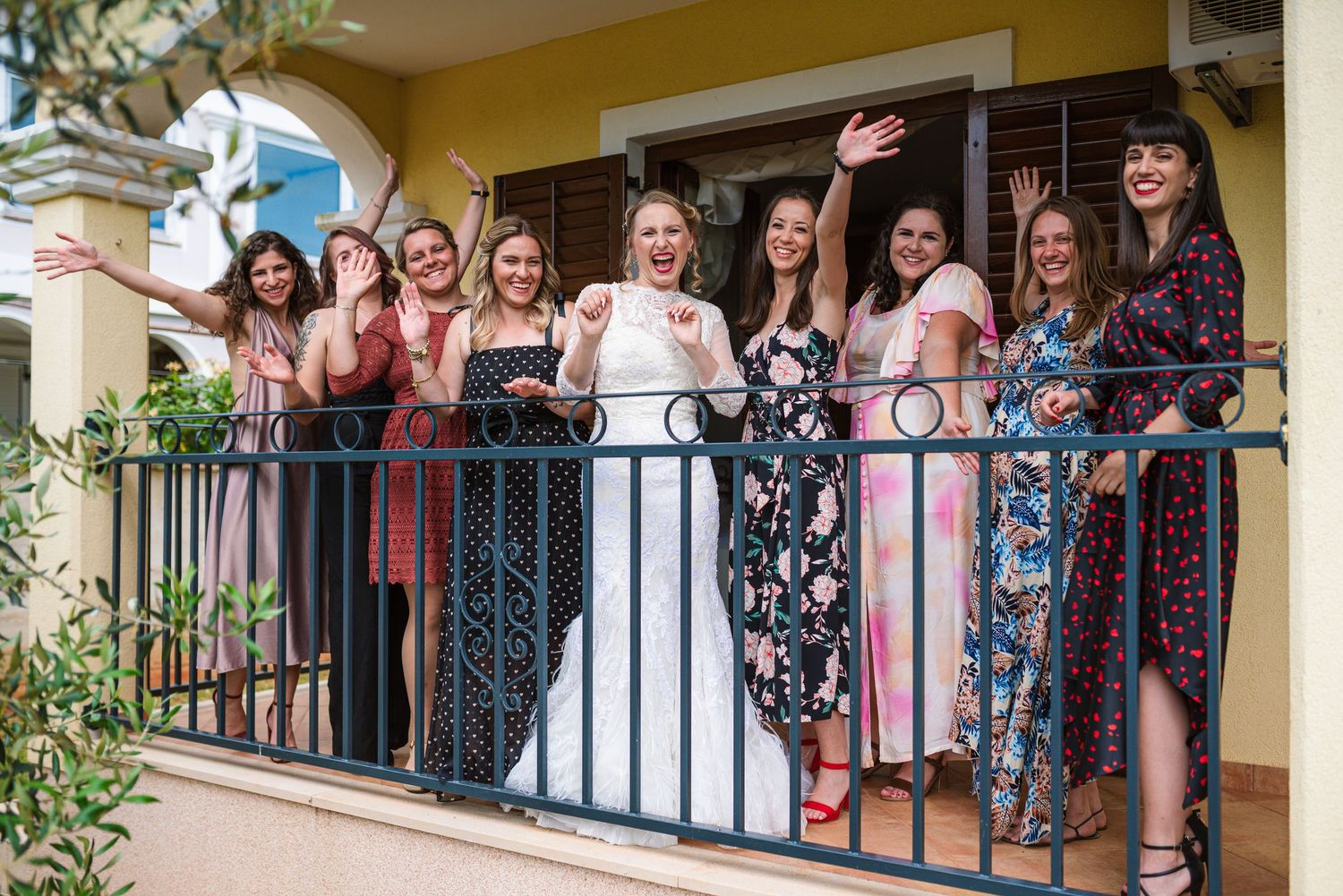 Bride and bridesmaids waving and cheering from a sunny balcony together.