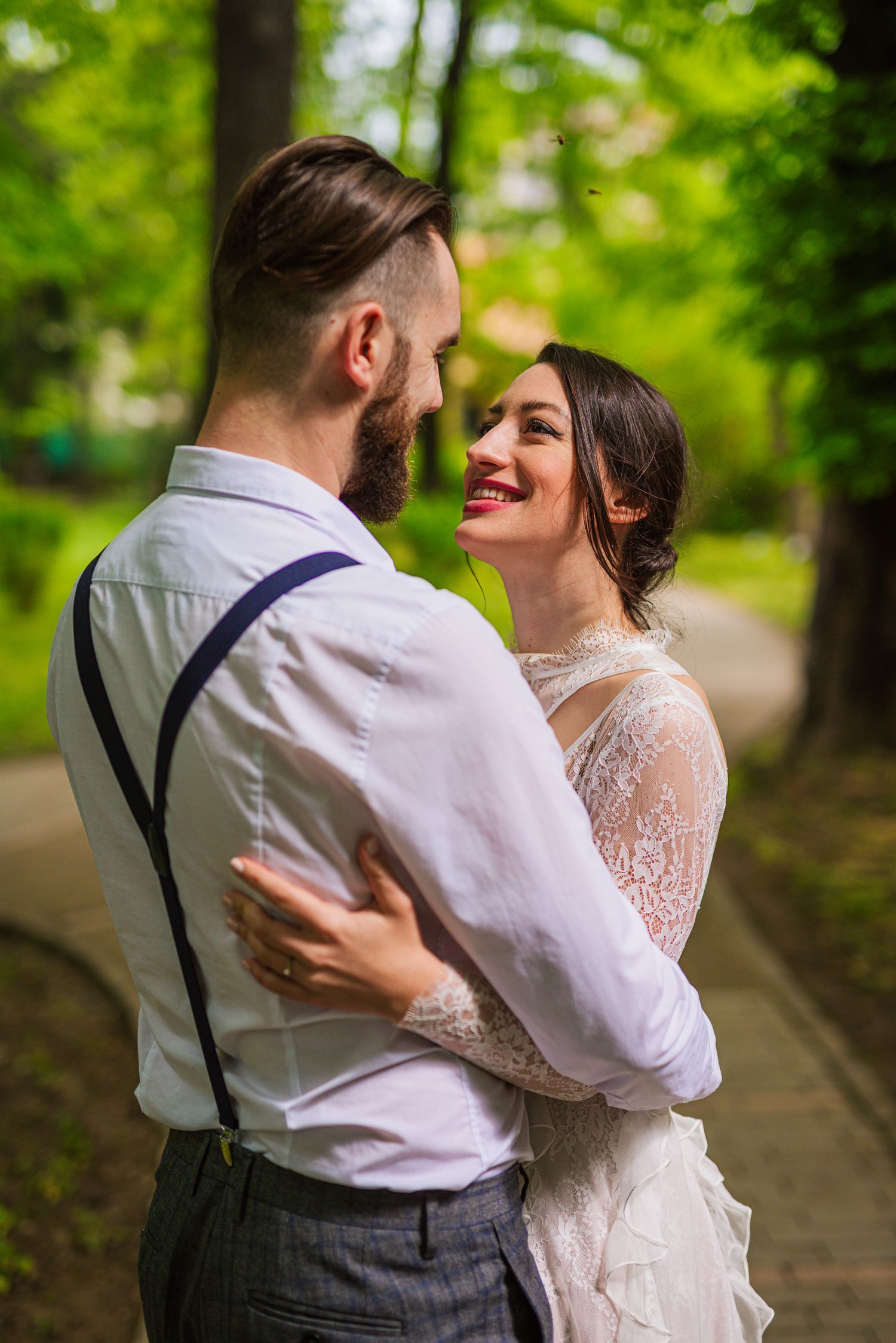 Smiling bride in lace wedding dress embraces bearded groom with suspenders in a green park.