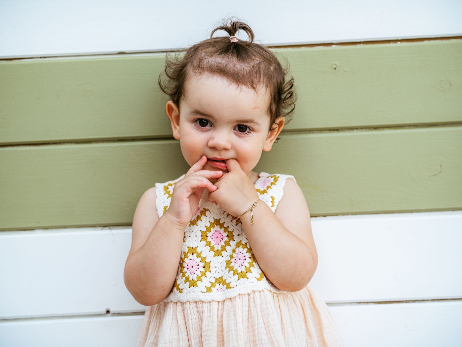 Adorable toddler girl in embroidered dress poses with hands near face against a green and white wooden wall.