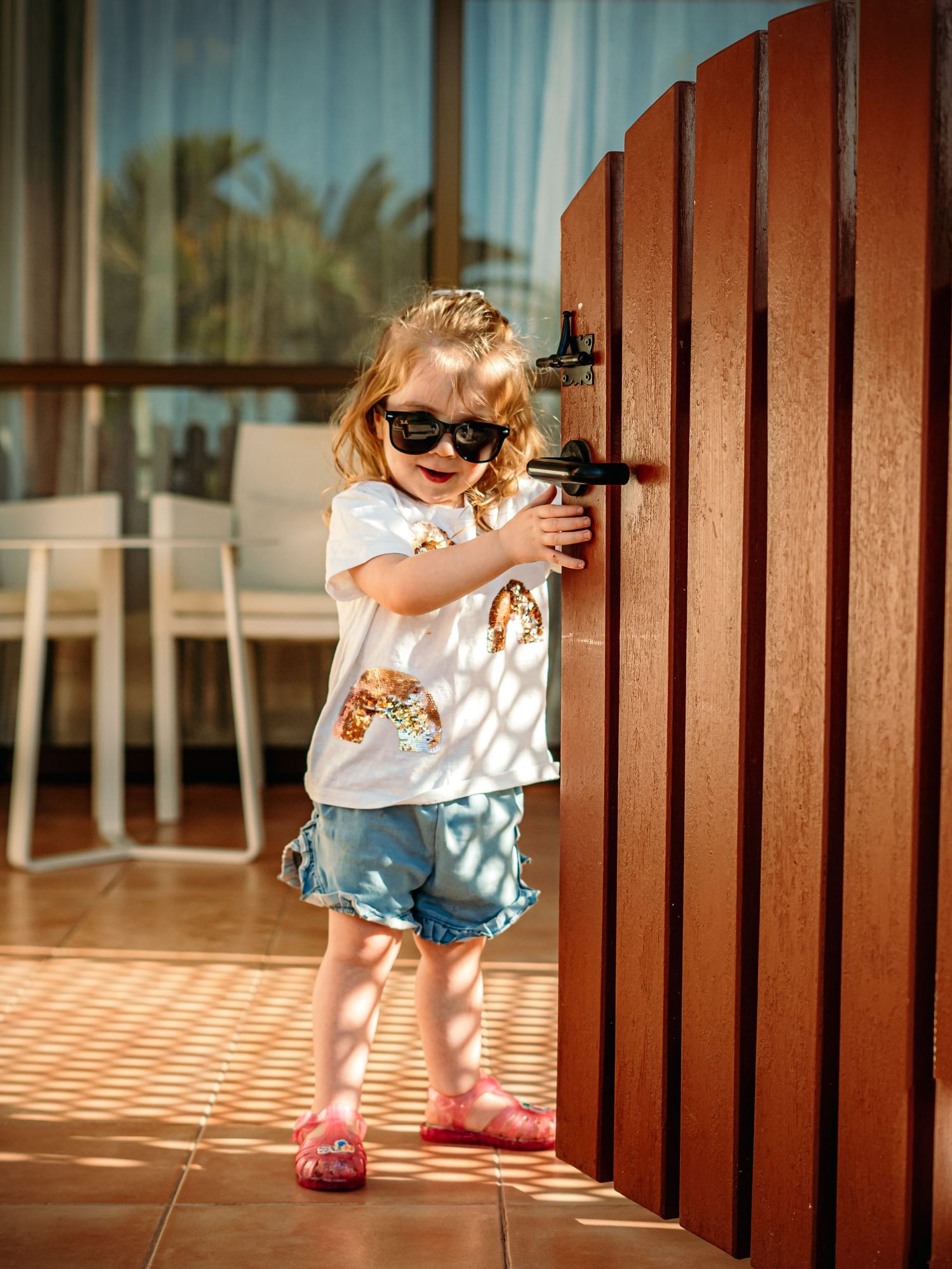 Cute blonde toddler girl wearing sunglasses and denim shorts opens a wooden gate on a sunny patio.