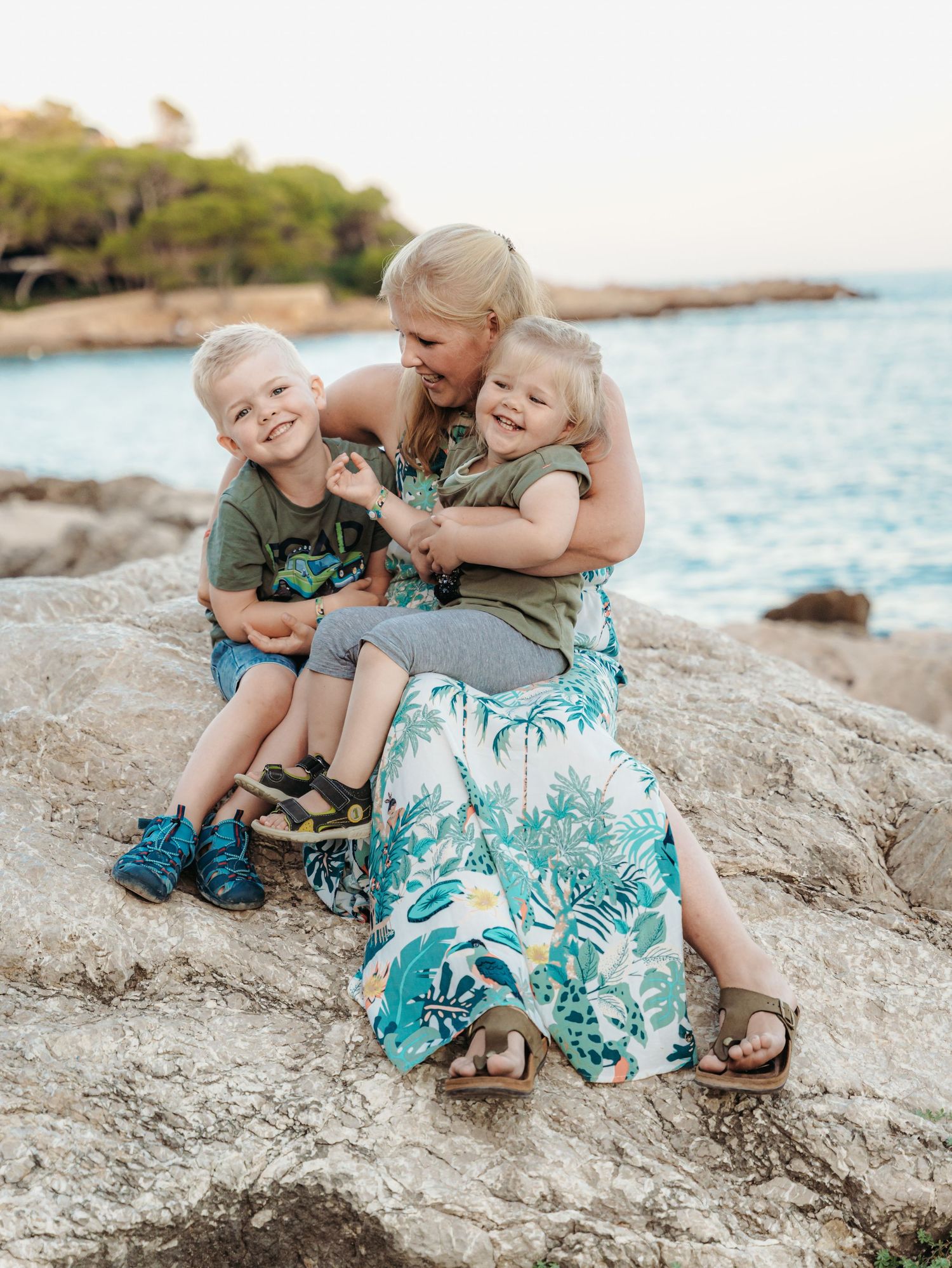 A blonde mother sits on coastal rocks hugging her two young children near a blue sea.