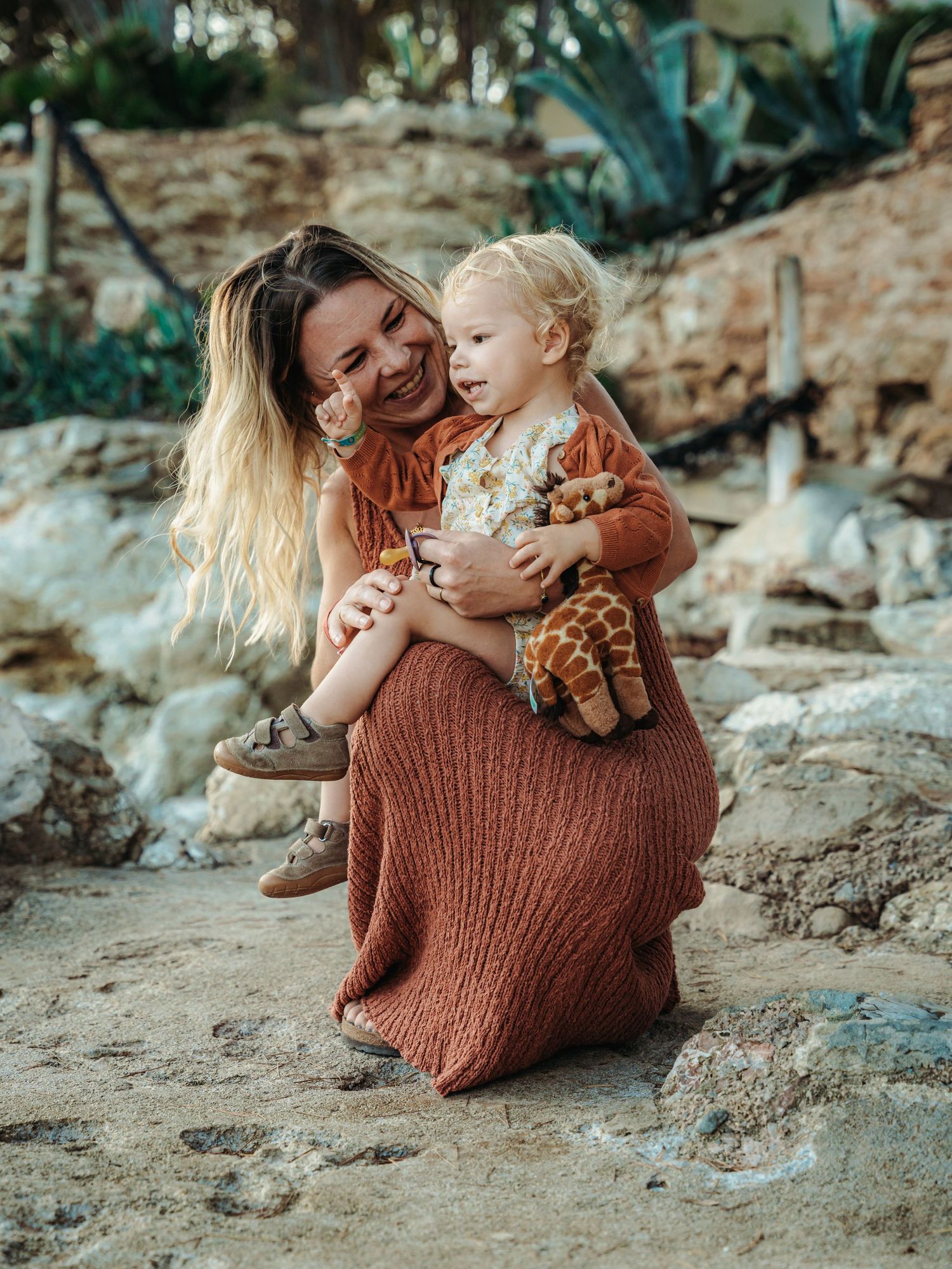 A joyful mother in a rust knit dress sits with her toddler clutching a giraffe plush toy outdoors.