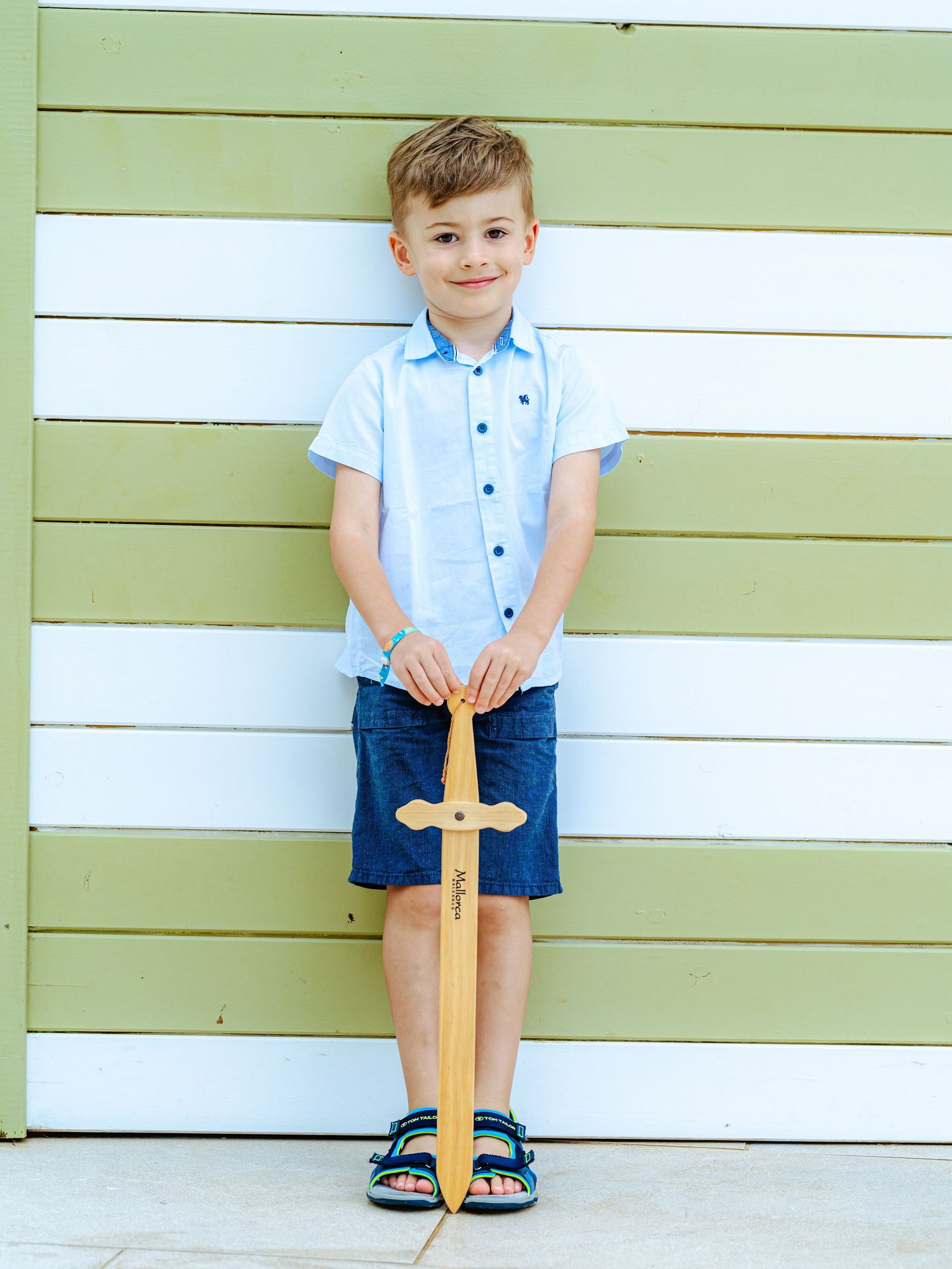 Young boy in light blue shirt smiling while holding a wooden toy sword against striped wall.