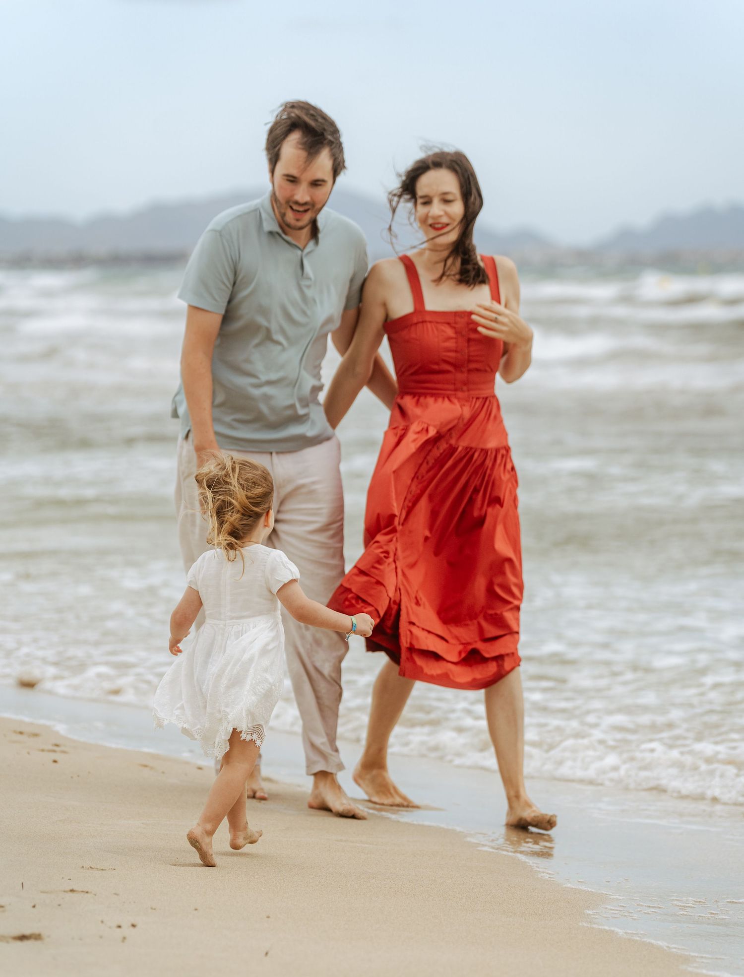 Happy family with young girl in white dress walking along a sunny beach with ocean waves nearby.