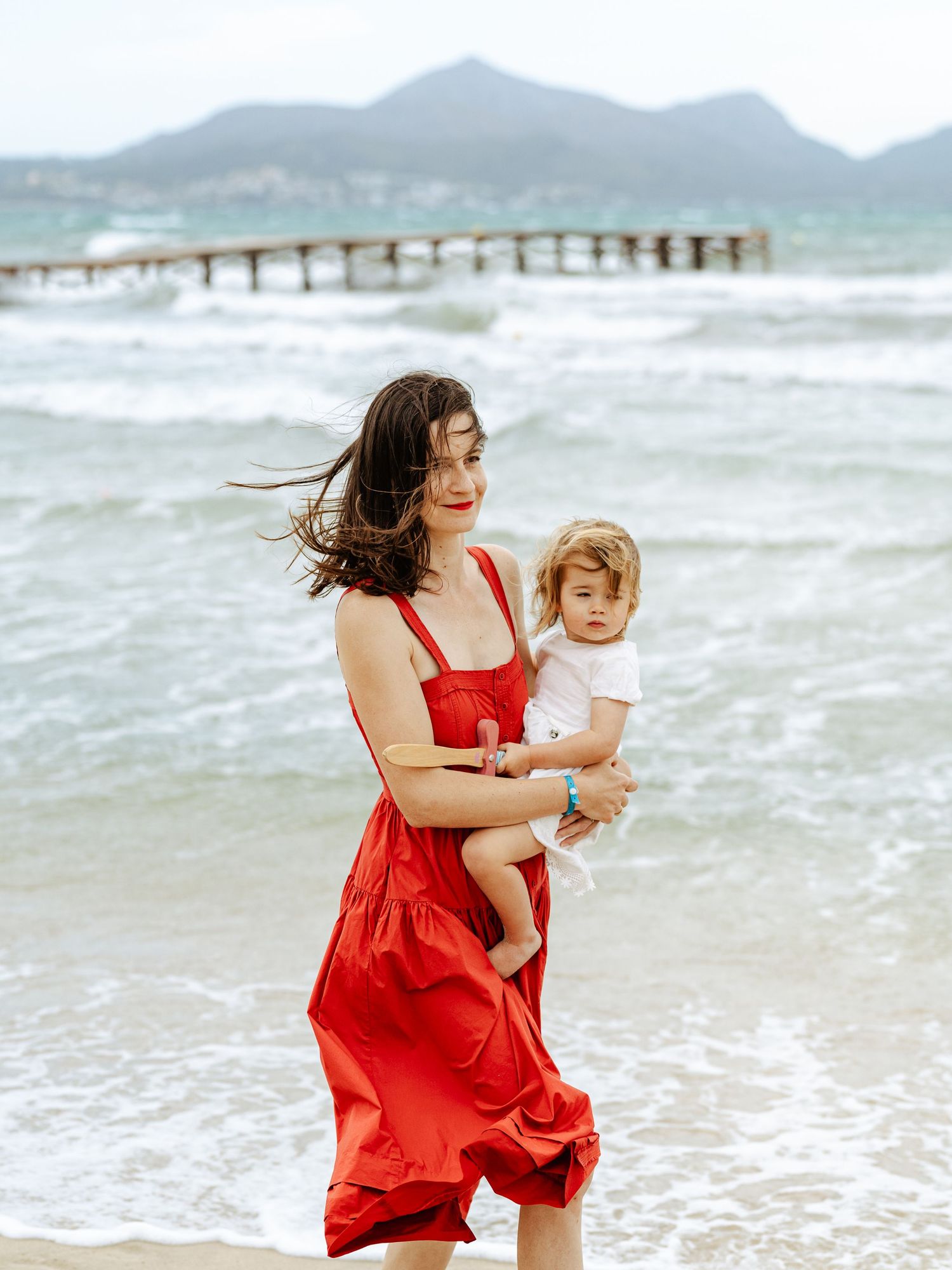 Mother in flowing red dress holding young child on windy beach with mountains in background.