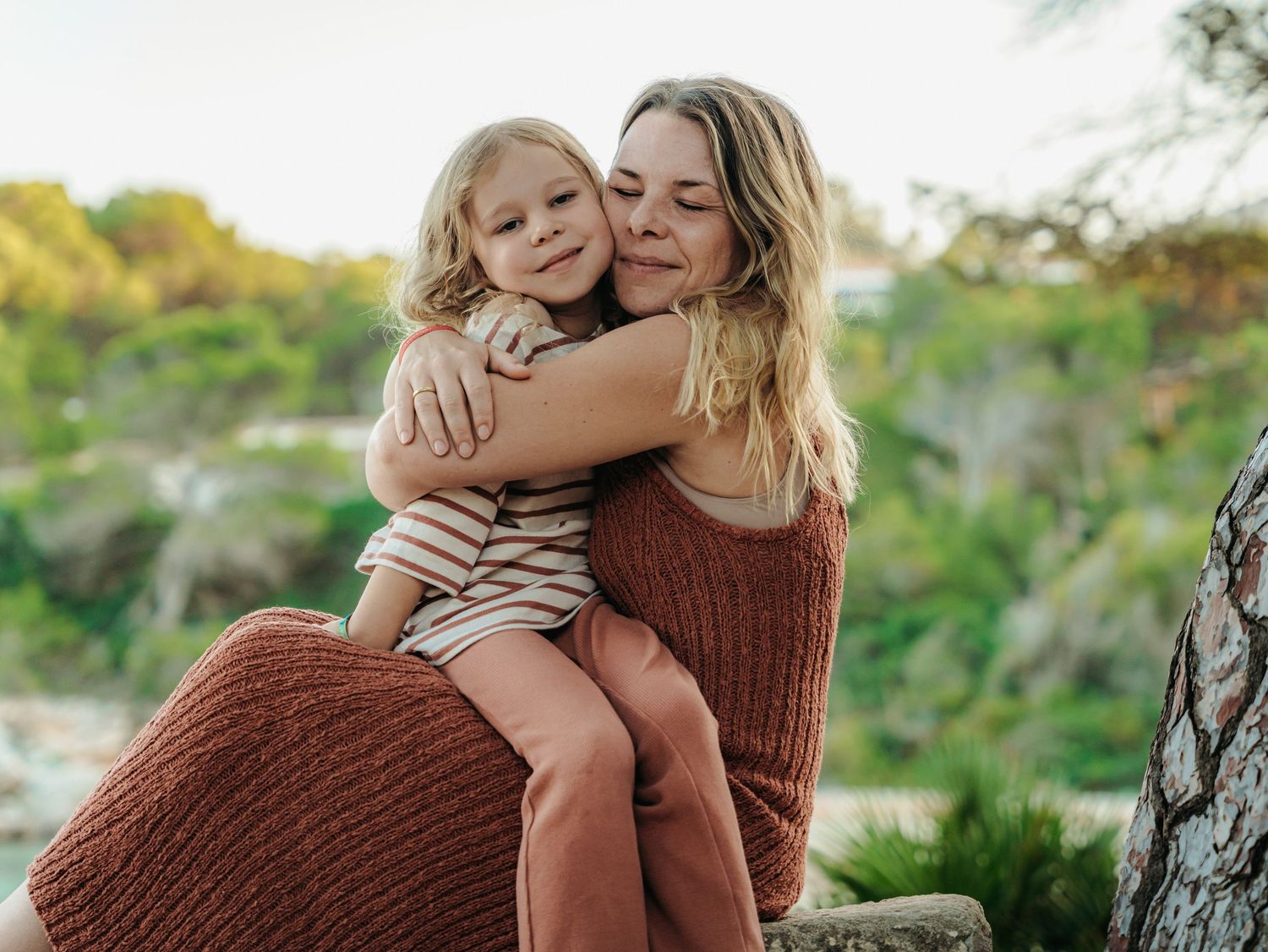 Mother and young daughter sharing a sweet hug in warm earth-toned outfits near coastal greenery.