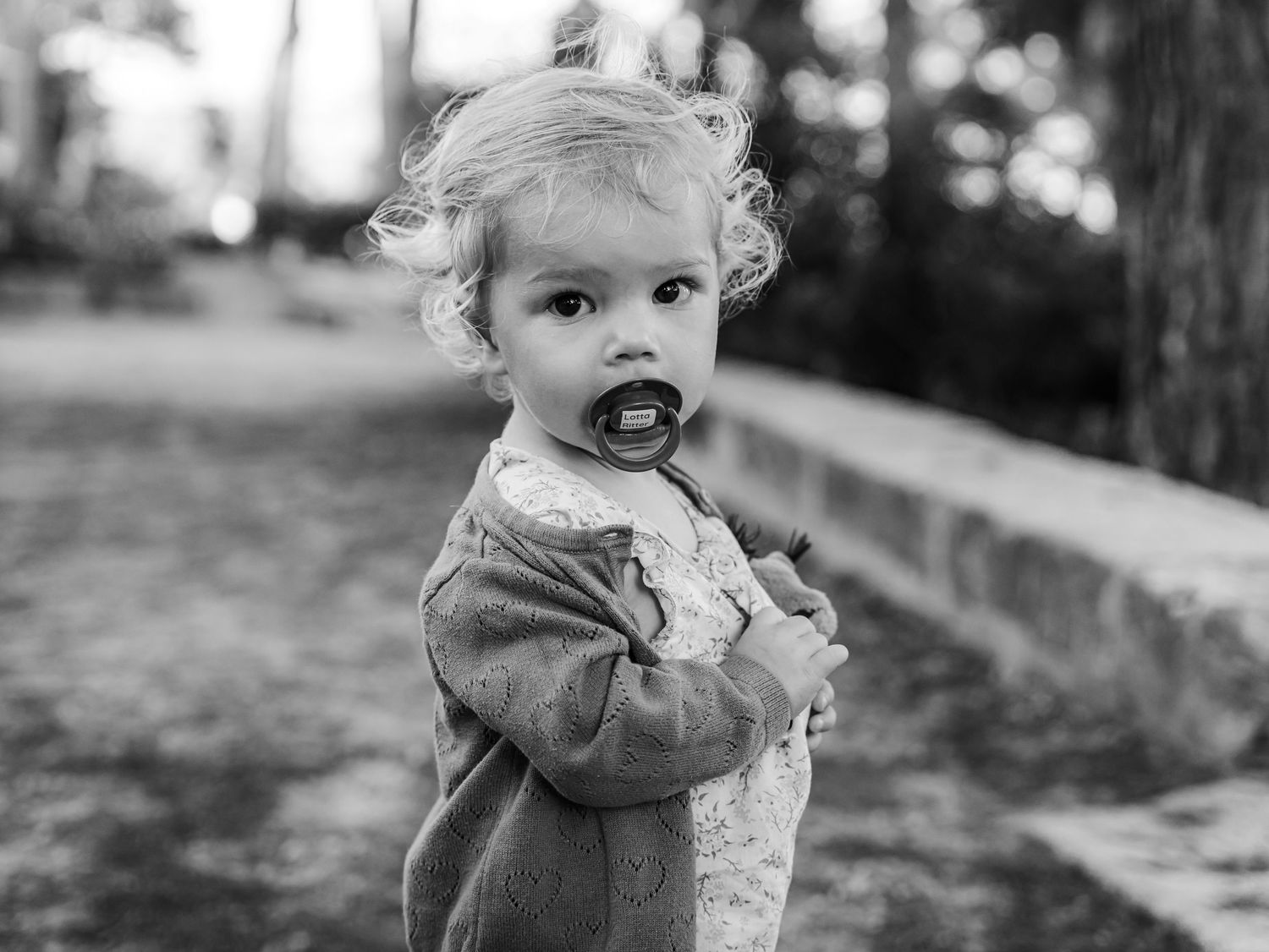 Black and white portrait of a cute toddler with a pacifier holding a stuffed toy outdoors.