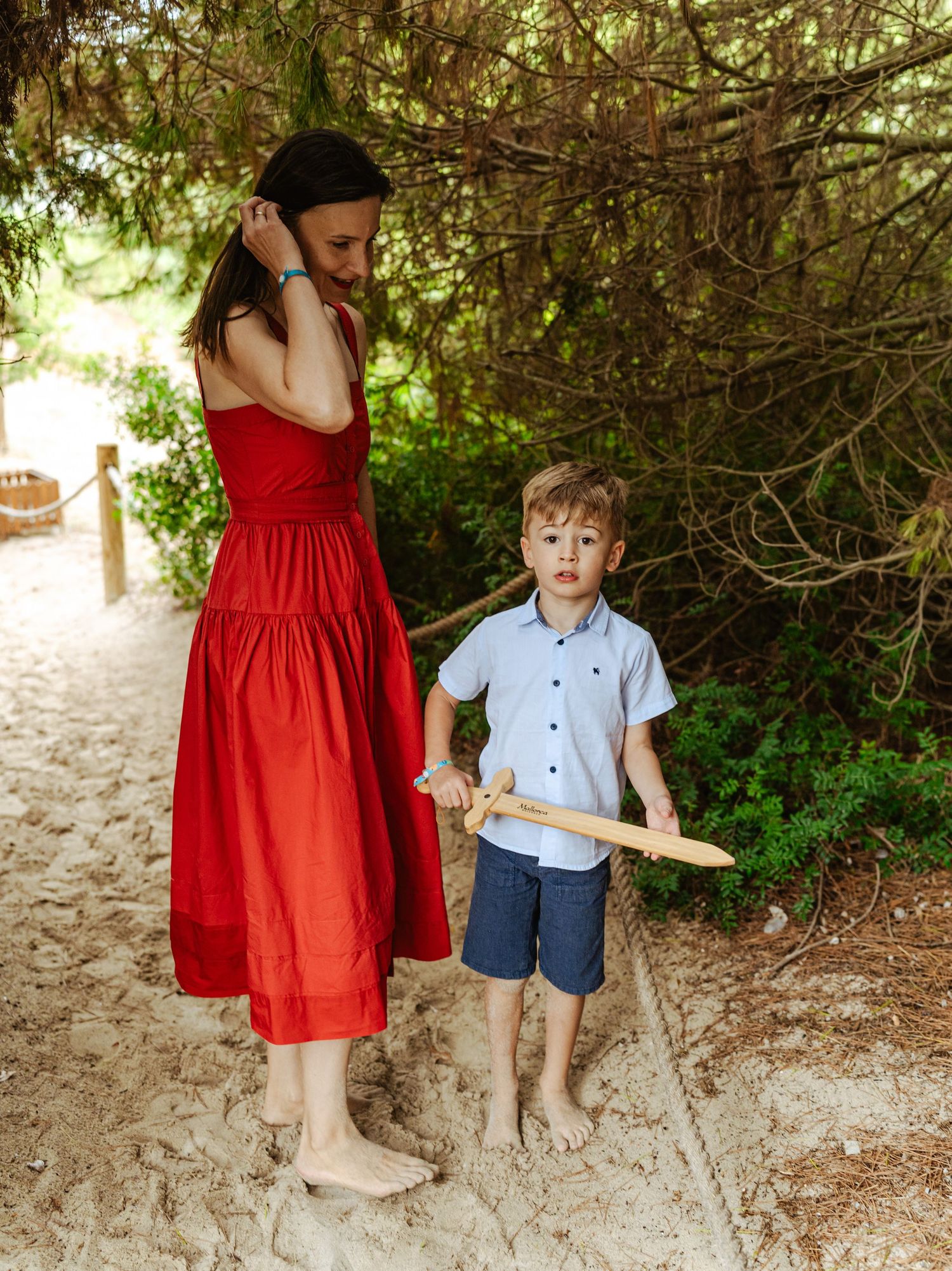 Mother in red dress standing with young boy holding wooden sword on sandy garden path.