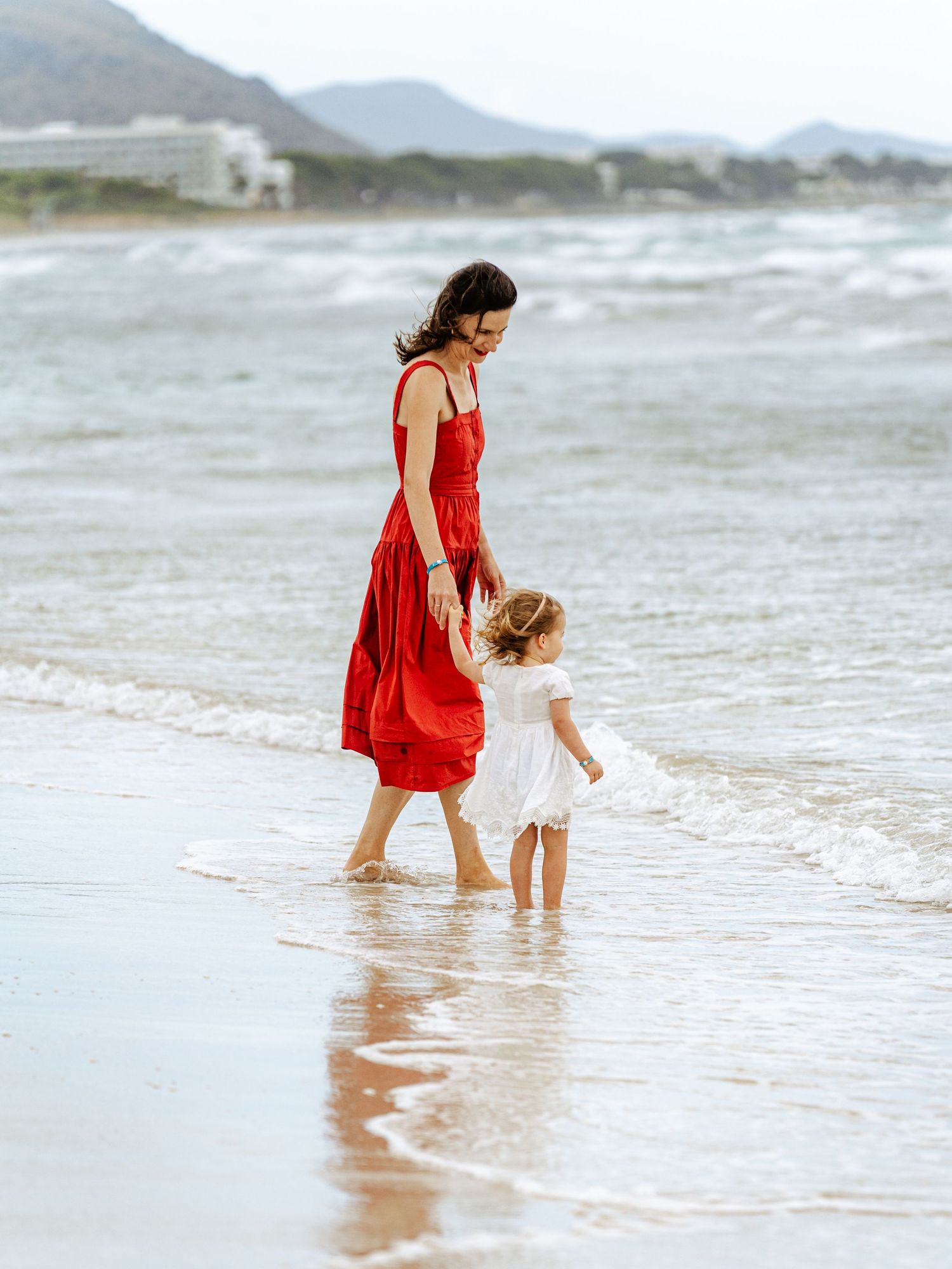 Mother wearing red sundress walks barefoot with toddler in white dress at the ocean's edge.