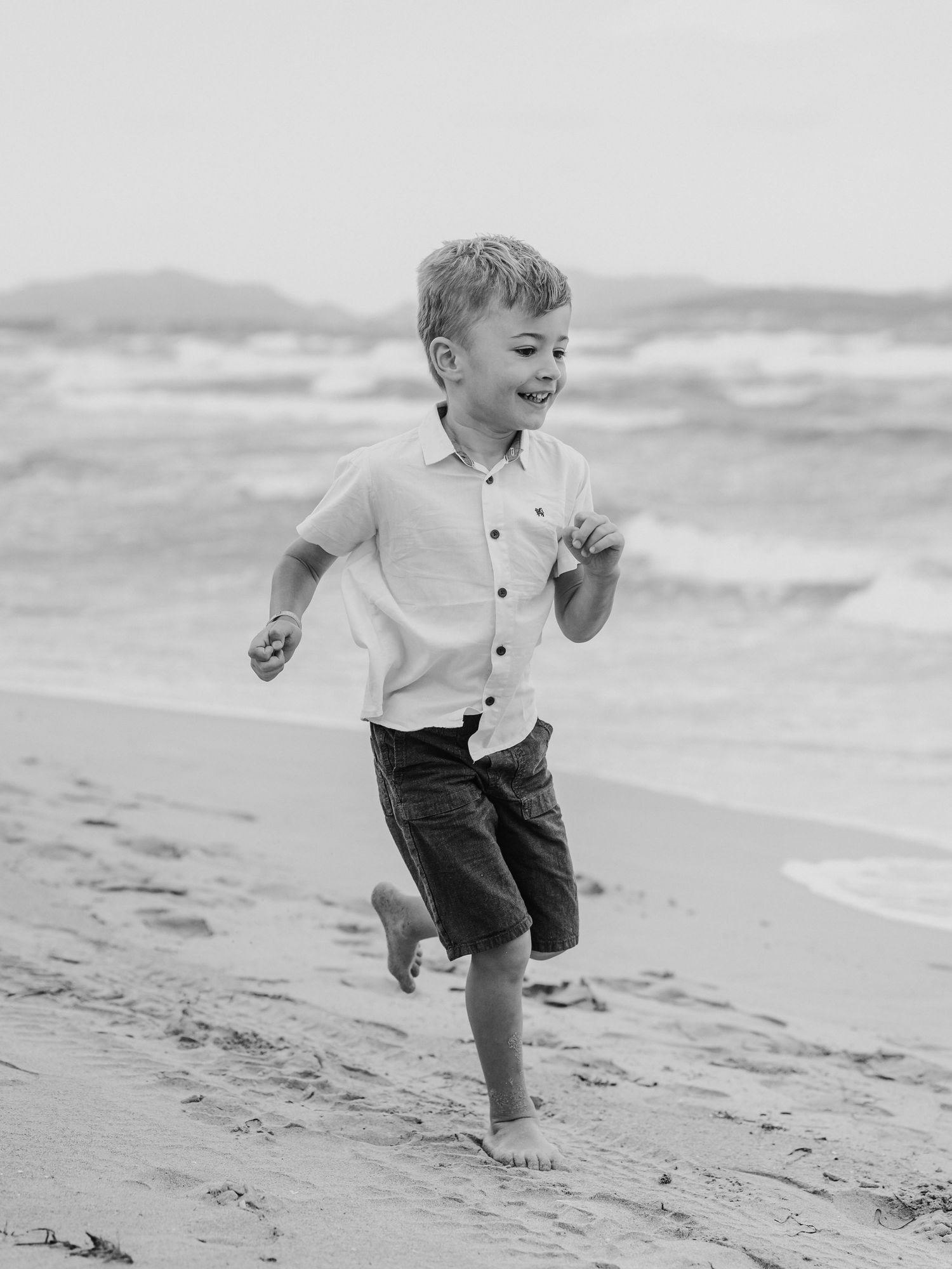 Black and white photo of a smiling boy running barefoot on a beach.