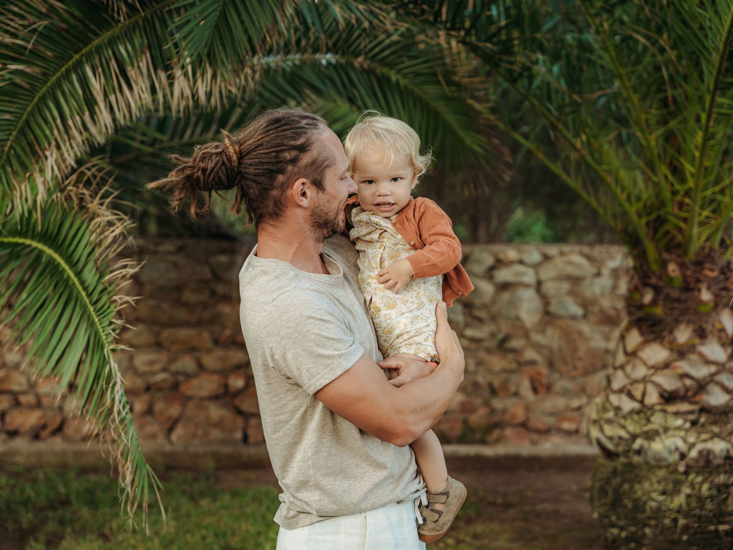 Young father with man bun tenderly holding toddler in his arms beside a lush palm tree.