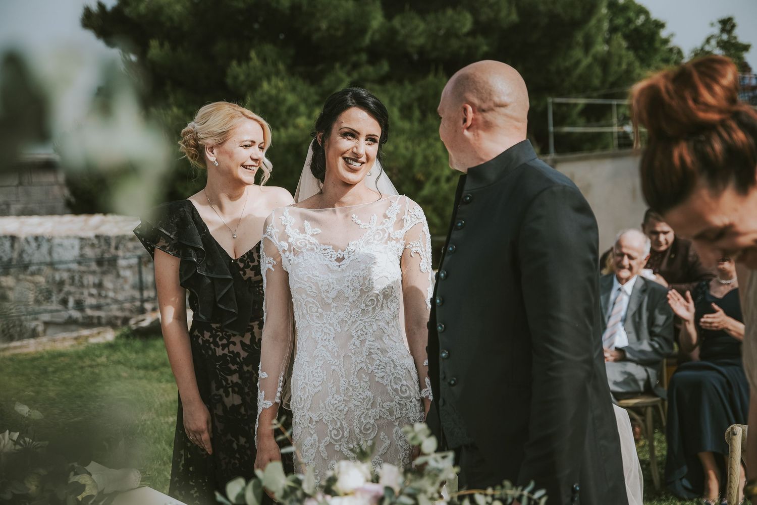 Smiling bride in lace wedding dress walking the aisle with her mother and father at outdoor ceremony.