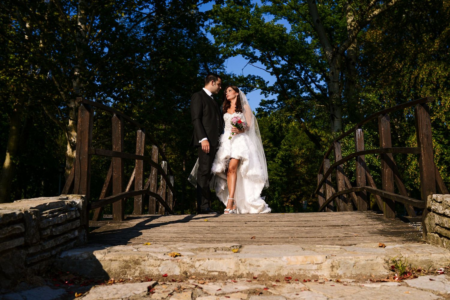 Bride and groom sharing a romantic moment on a rustic stone bridge surrounded by lush green trees on their wedding day.