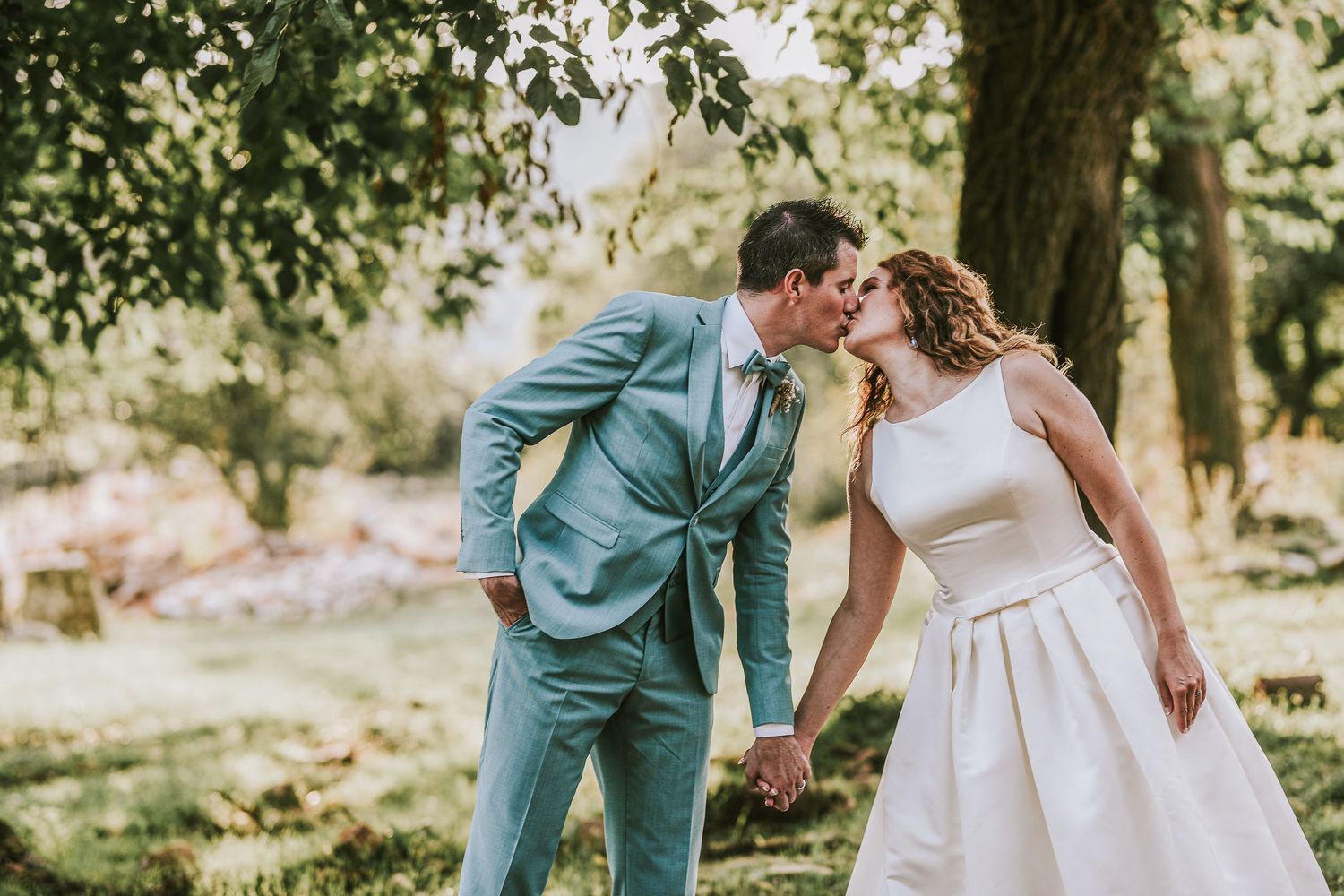 Bride and groom kissing while holding hands in a sunlit green garden setting.