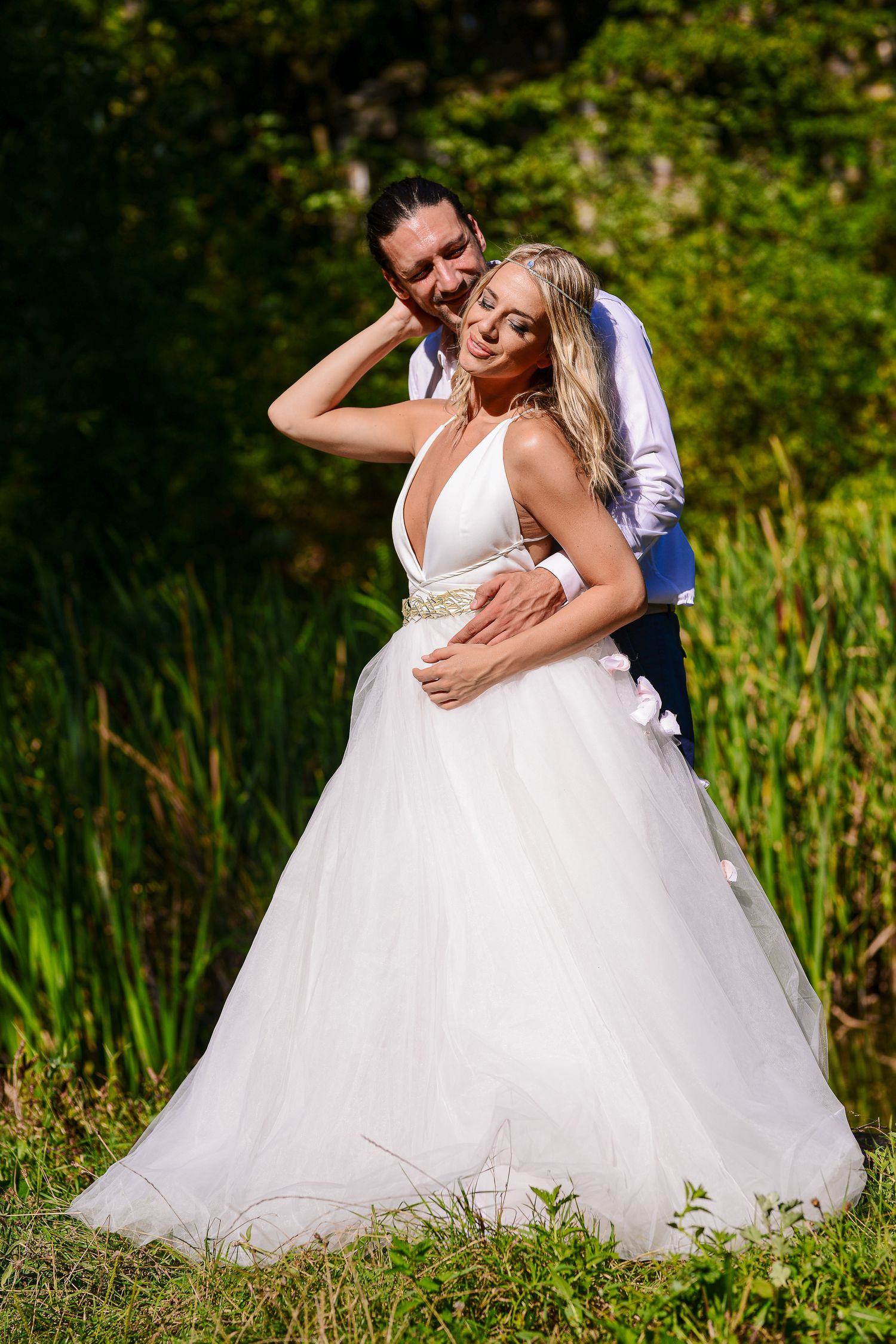 Groom tenderly embracing bride in a white tulle gown beside a lush green pond in sunlight.