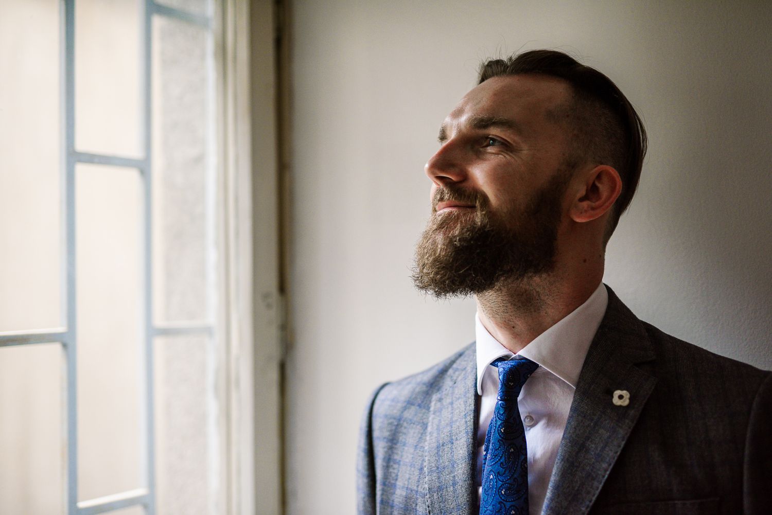 Handsome bearded groom in gray suit smiling joyfully while standing near a bright window.