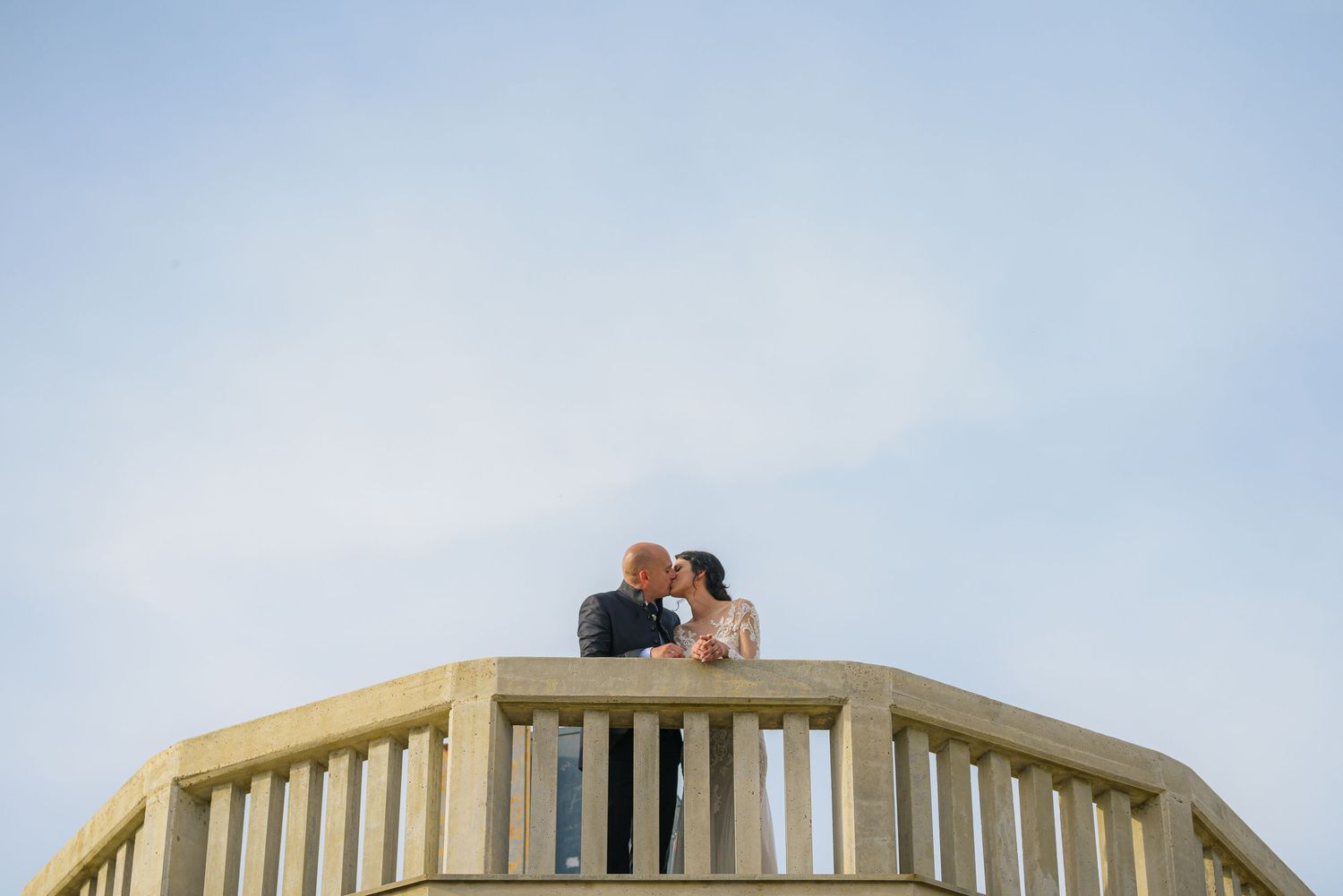 Bride and groom share a kiss atop a wooden balcony structure against a soft blue sky.