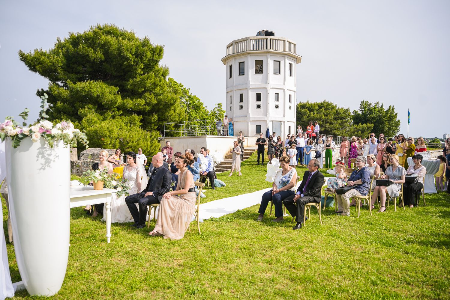 Outdoor wedding ceremony with seated guests on lawn near white tower landmark venue.