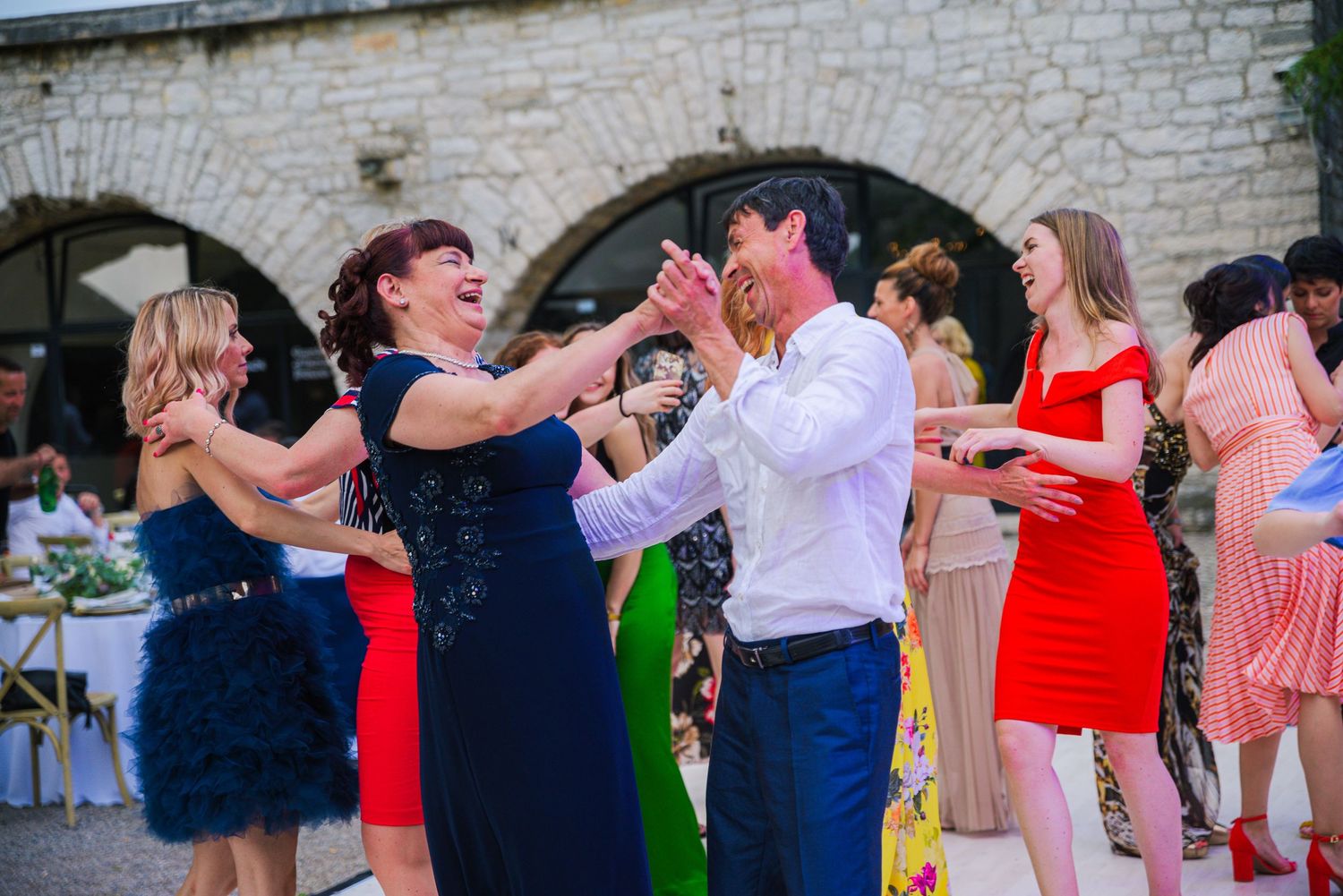 Wedding guests laugh and dance together in colorful attire at an outdoor reception.