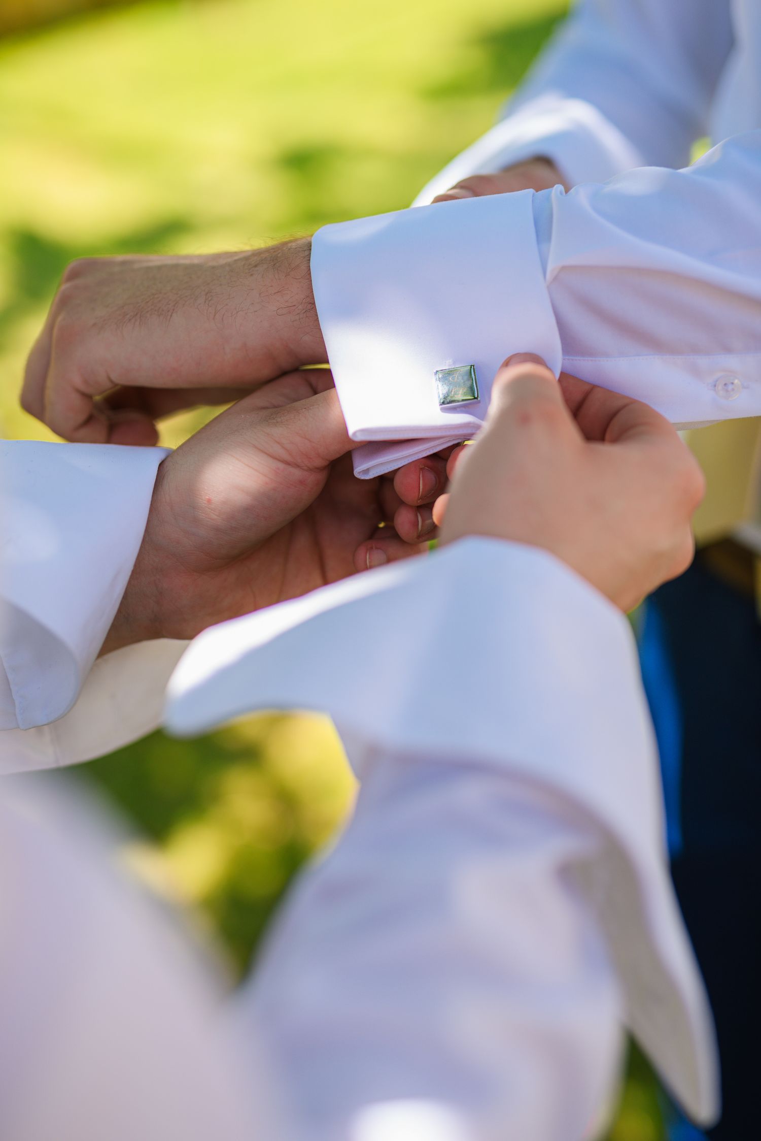 Best man fastens silver cufflinks on groom's white dress shirt sleeve outdoors.