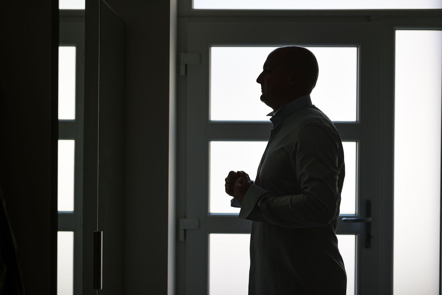 Groom silhouette standing by window adjusting cufflinks before wedding ceremony.