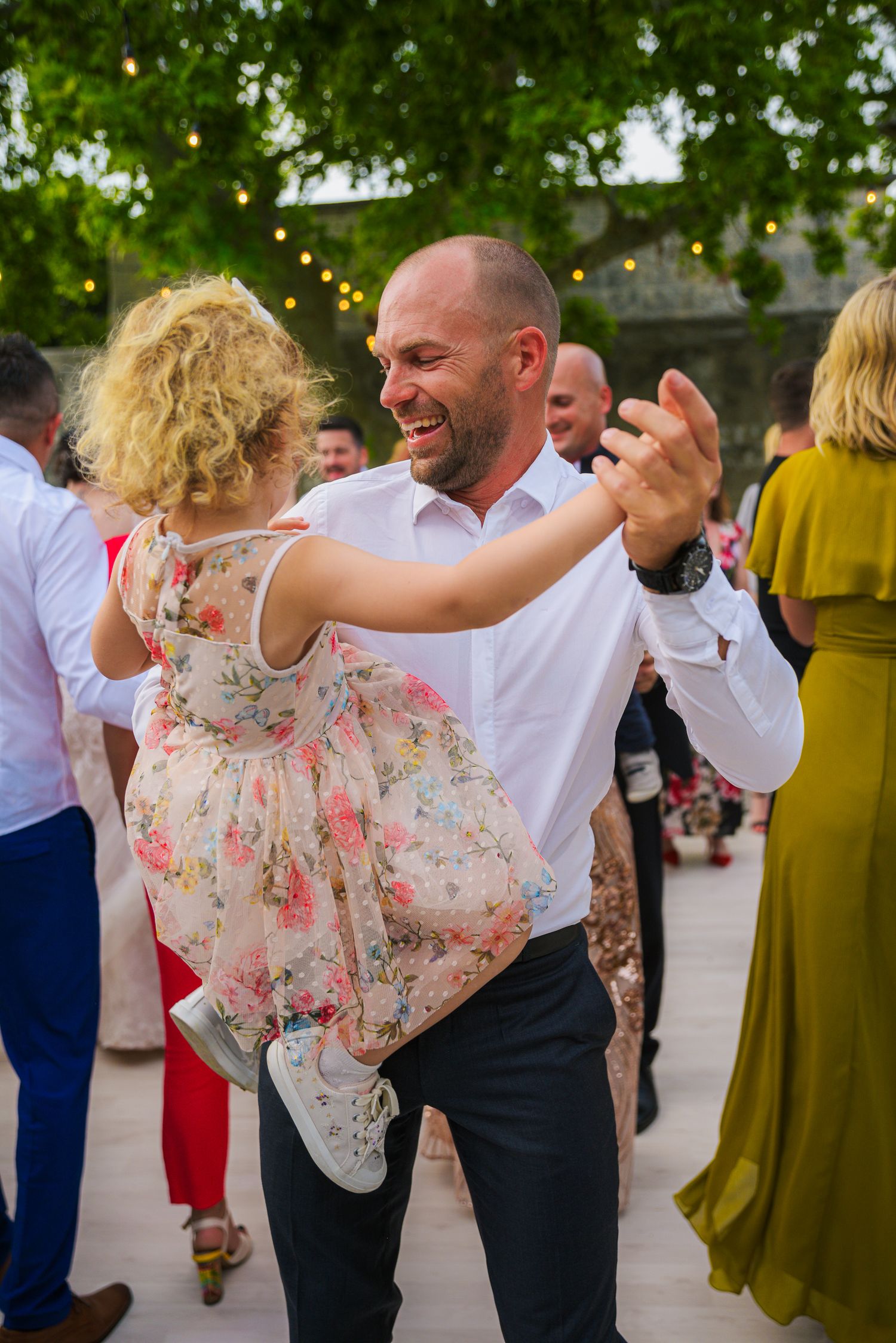 Joyful man in white shirt dances with a young girl in floral dress at wedding reception.