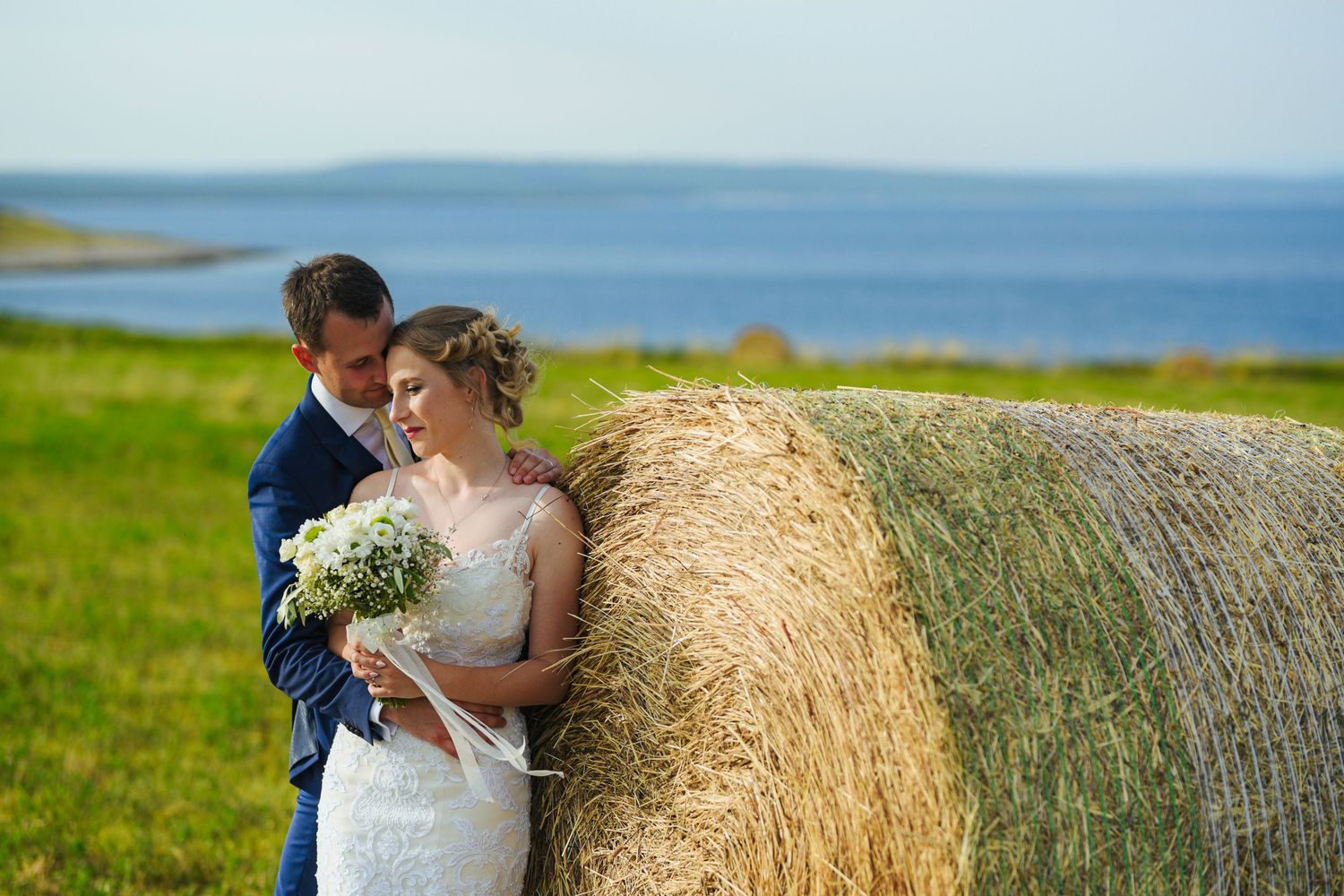 Romantic bride and groom lean against a hay bale with stunning ocean views in background.