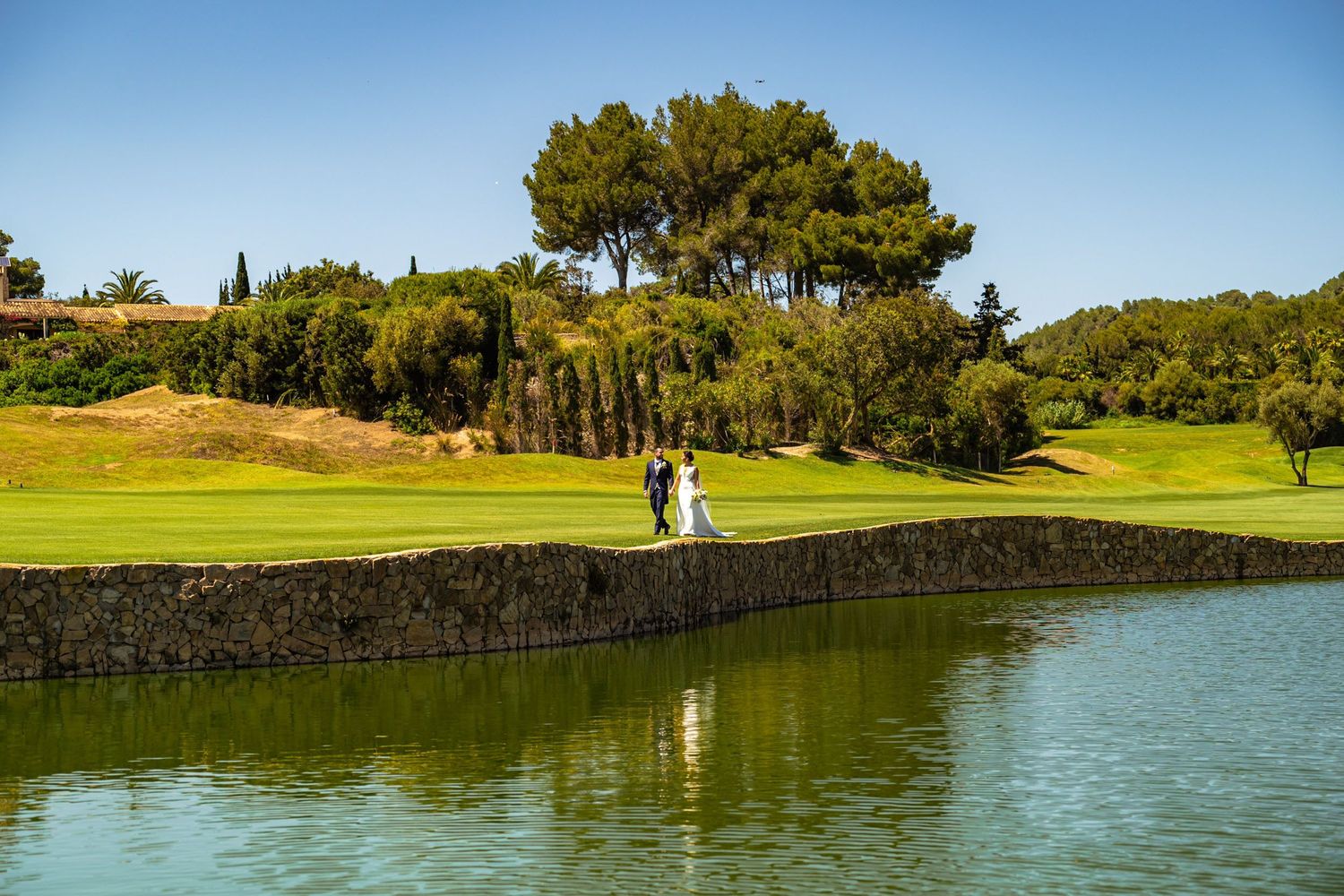 Bride and groom pose together on stone bridge over reflective pond at golf course venue.