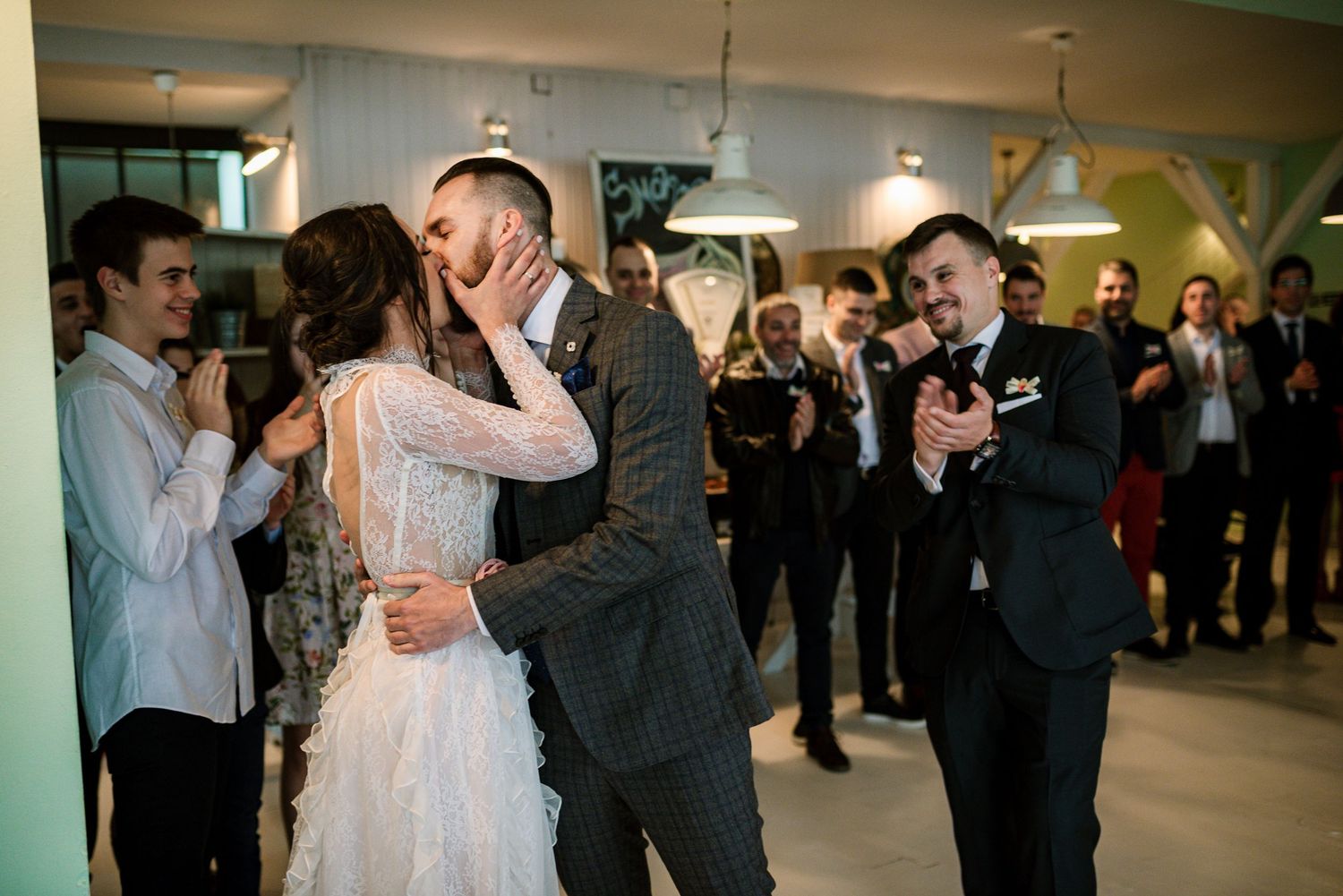 Bride and groom share passionate kiss while guests clap and celebrate at wedding reception.