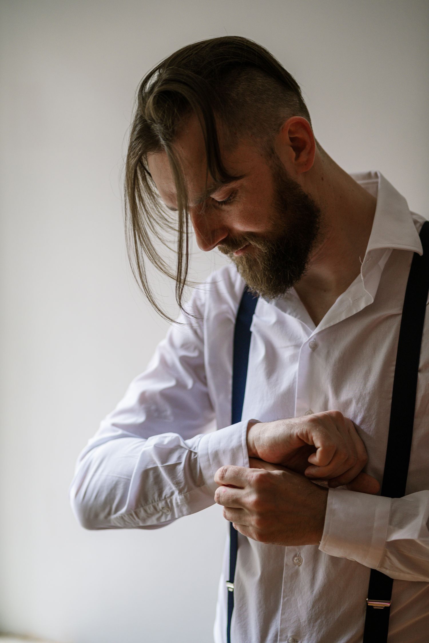 Smiling bearded groom in white shirt and suspenders fastening his cufflinks.