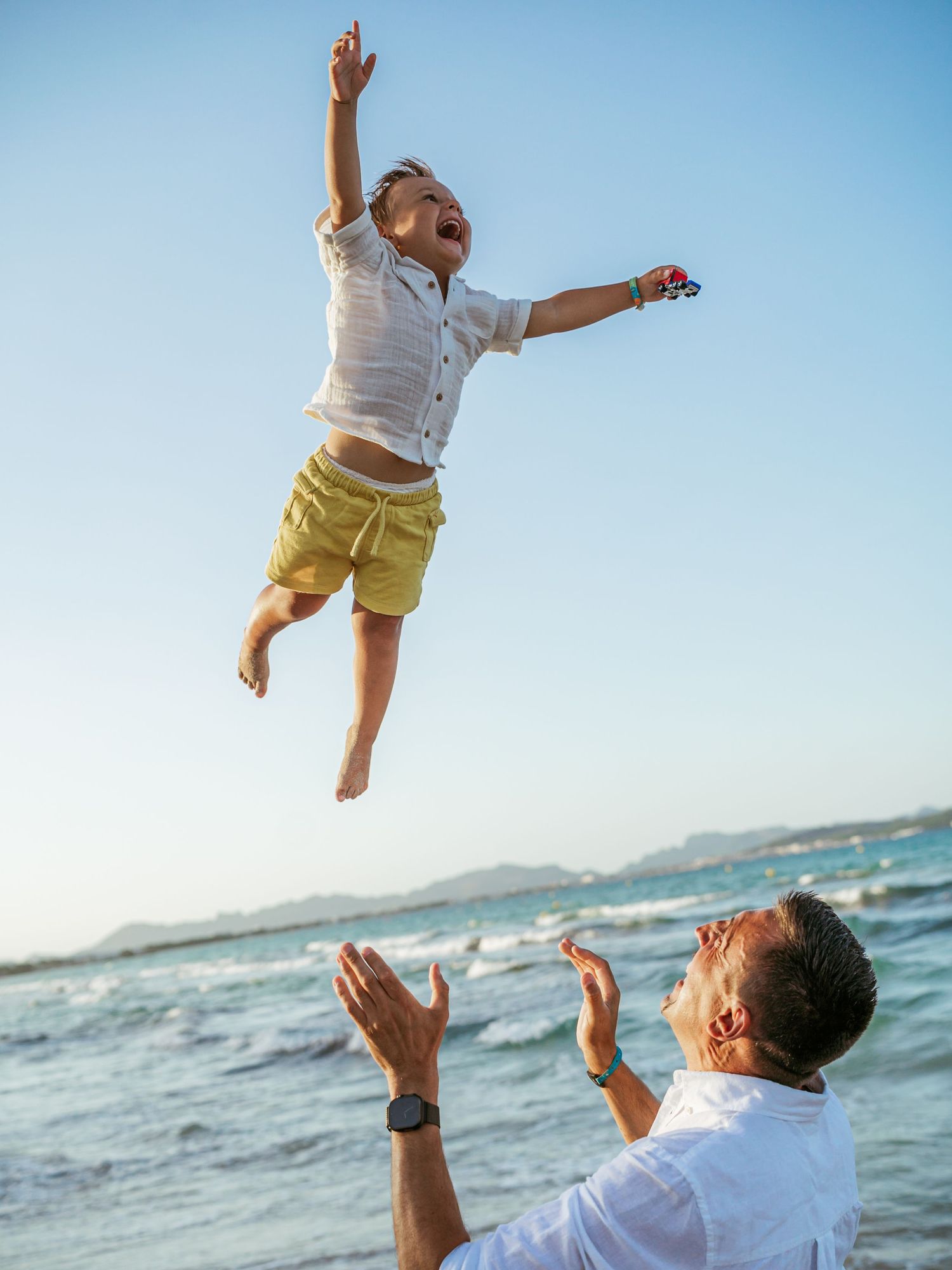 Excited young boy in yellow shorts leaping joyfully into the air above his fathers outstretched hands.