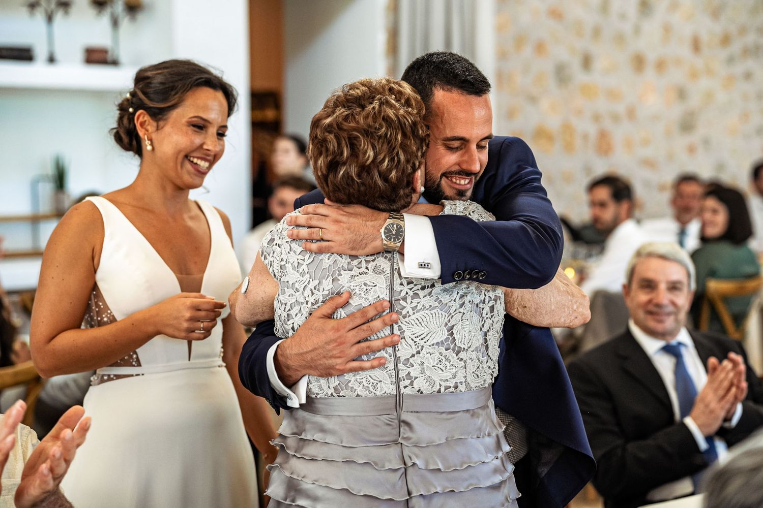 Groom embraces a family member warmly as smiling bride watches during a heartfelt wedding ceremony moment.