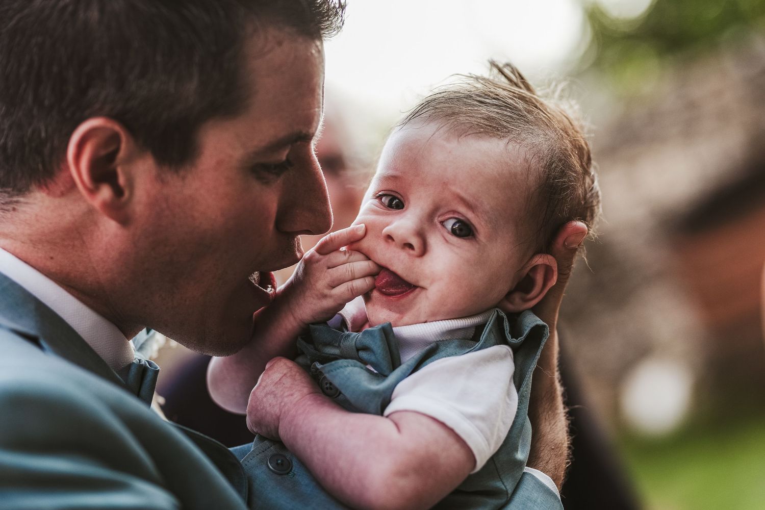 Groom in blue suit tenderly holding a smiling baby at an outdoor wedding celebration.