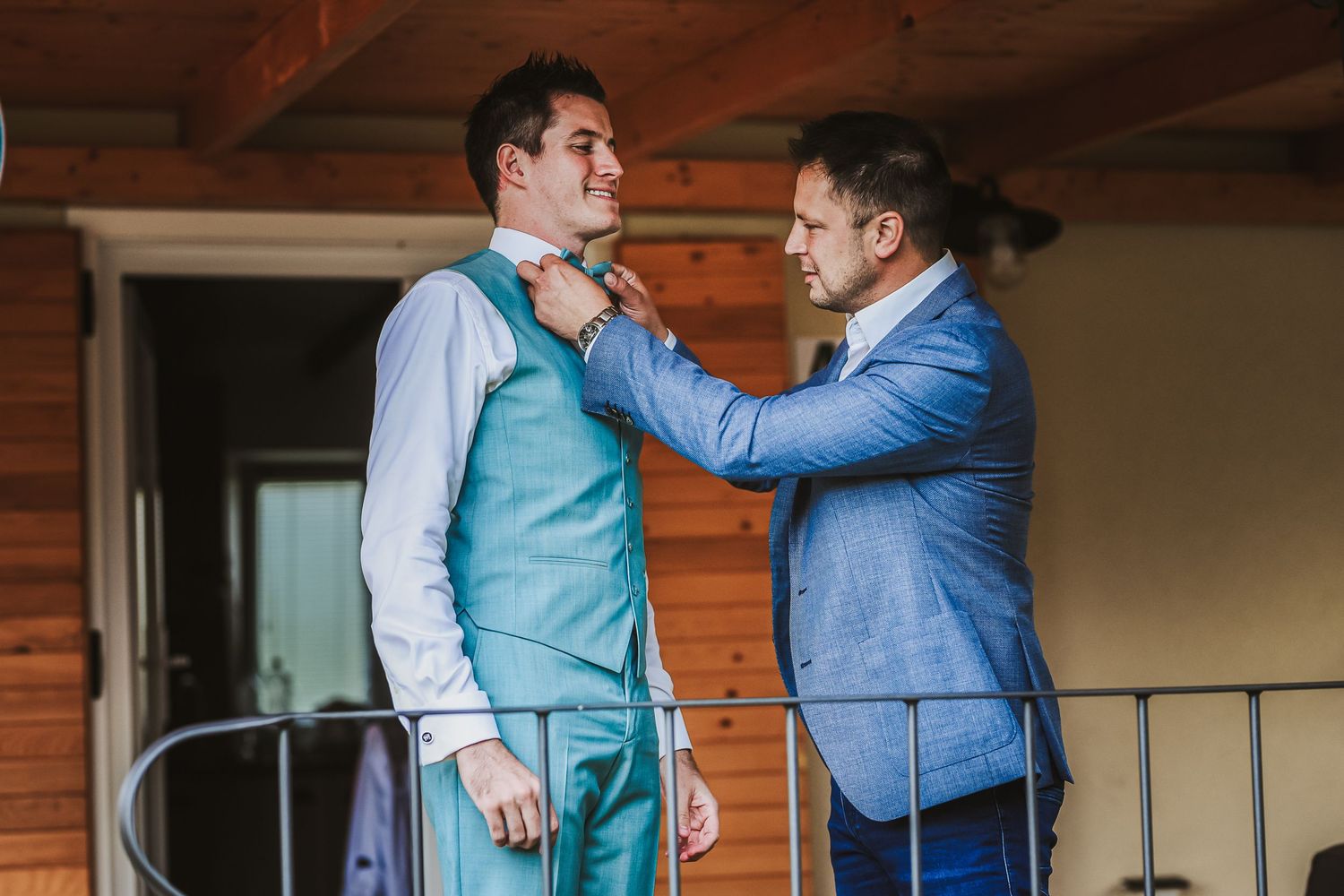 Groomsman helping groom adjust his teal bow tie on a wooden balcony before the wedding ceremony.