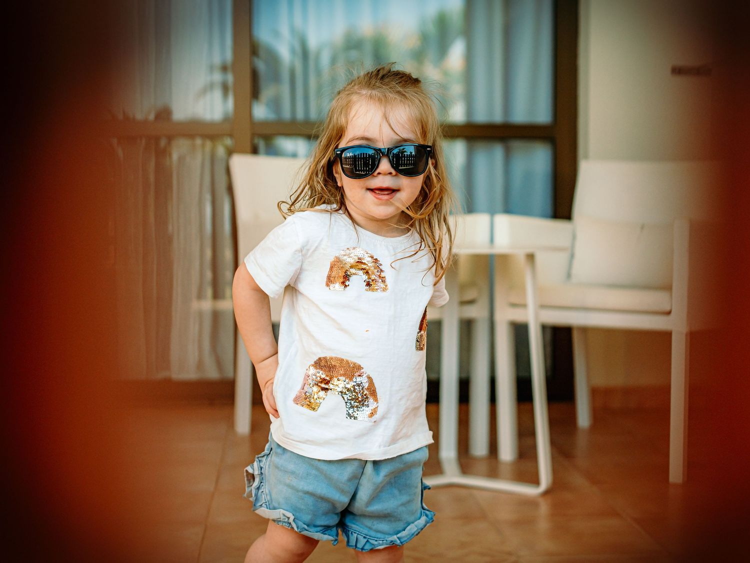 Stylish toddler girl wearing sunglasses and sequin t-shirt with blue denim shorts indoors.