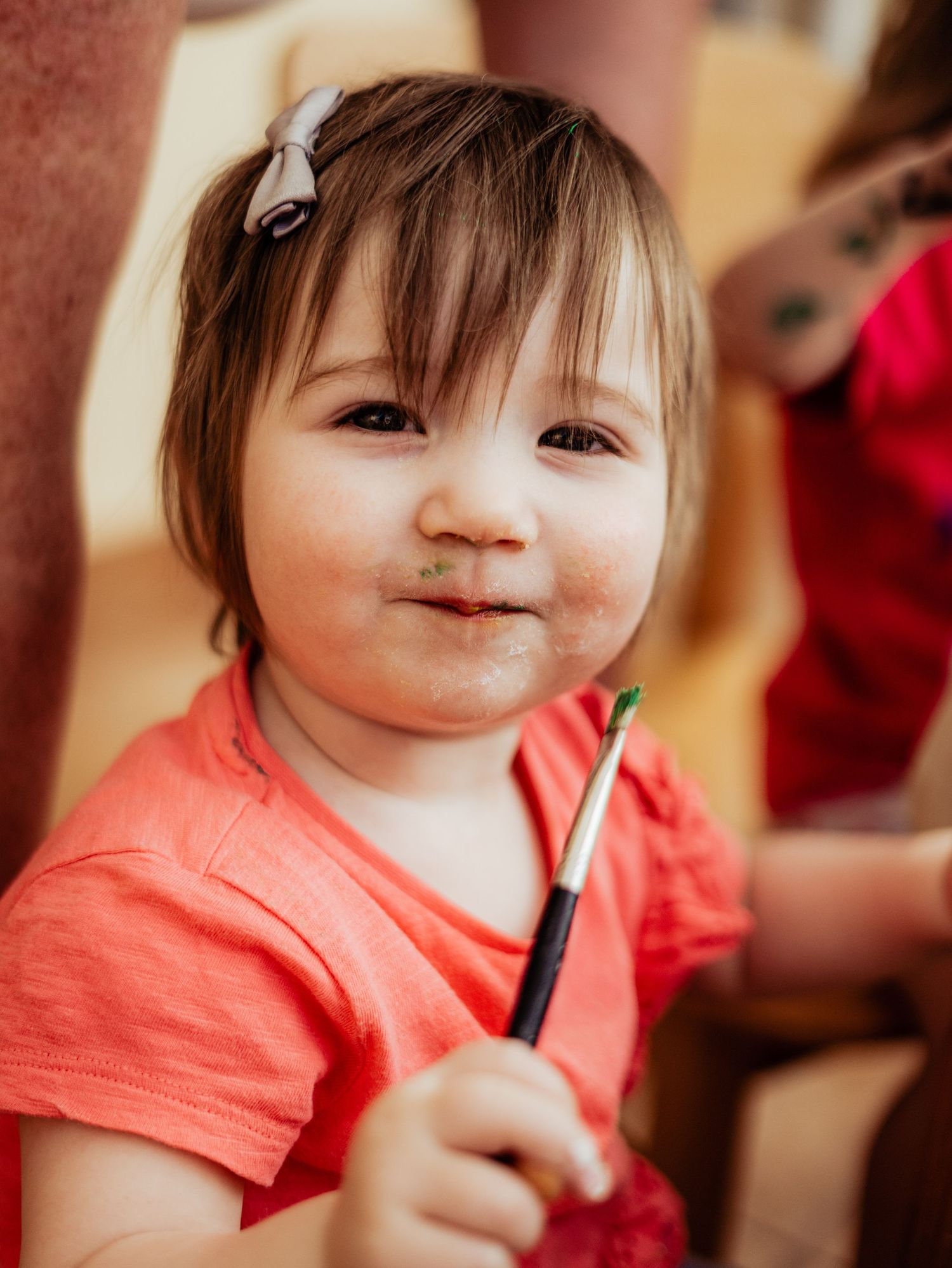 An adorable toddler girl smiles at the camera while holding a green paintbrush during an art activity.