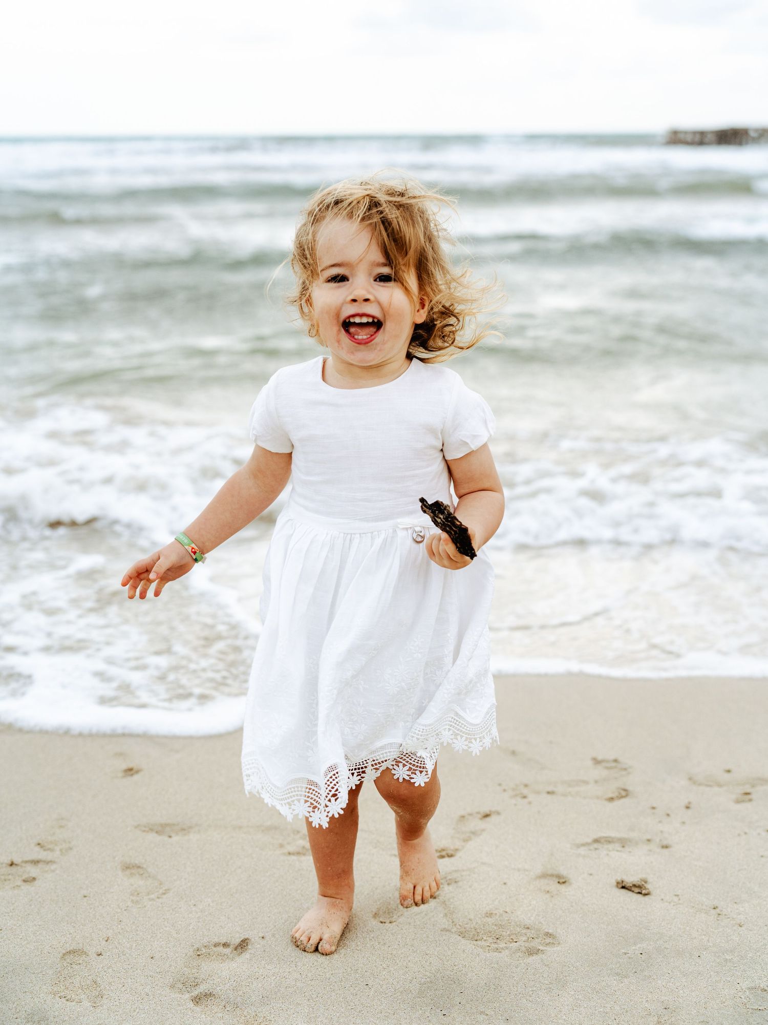 Happy laughing toddler girl in white lace dress playing barefoot on a sandy beach.