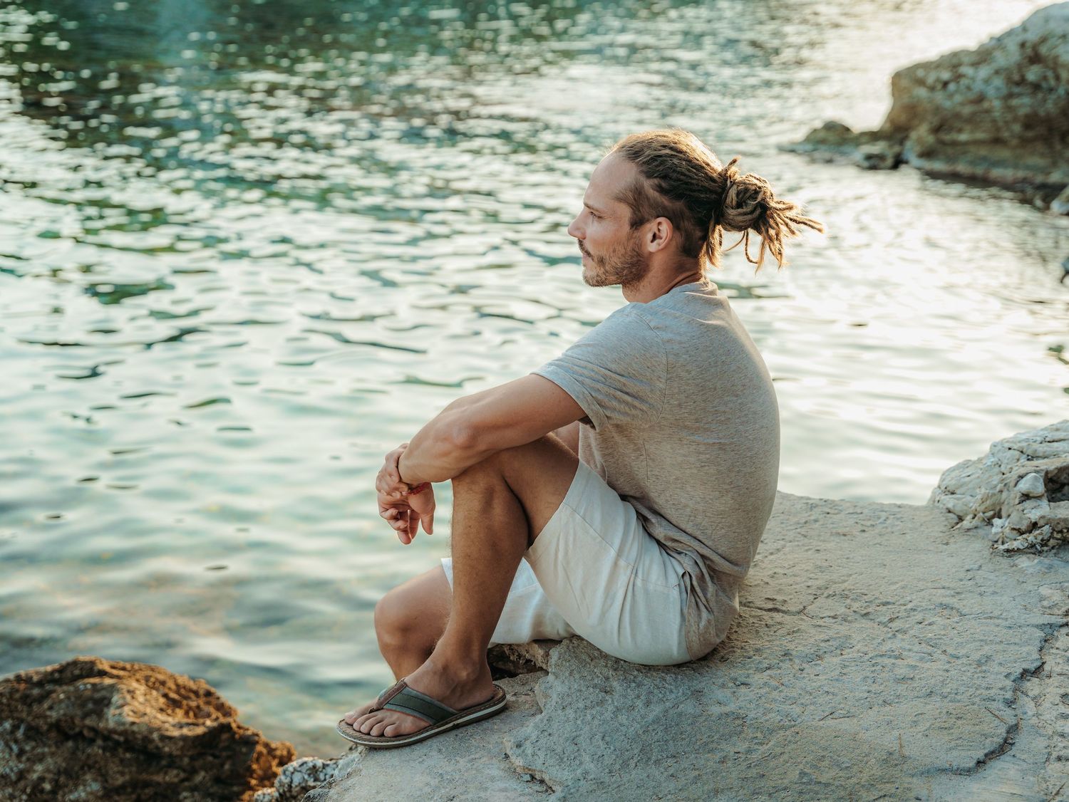 A man with tied-back hair sits peacefully on rocks beside shimmering water in a gray shirt and shorts.
