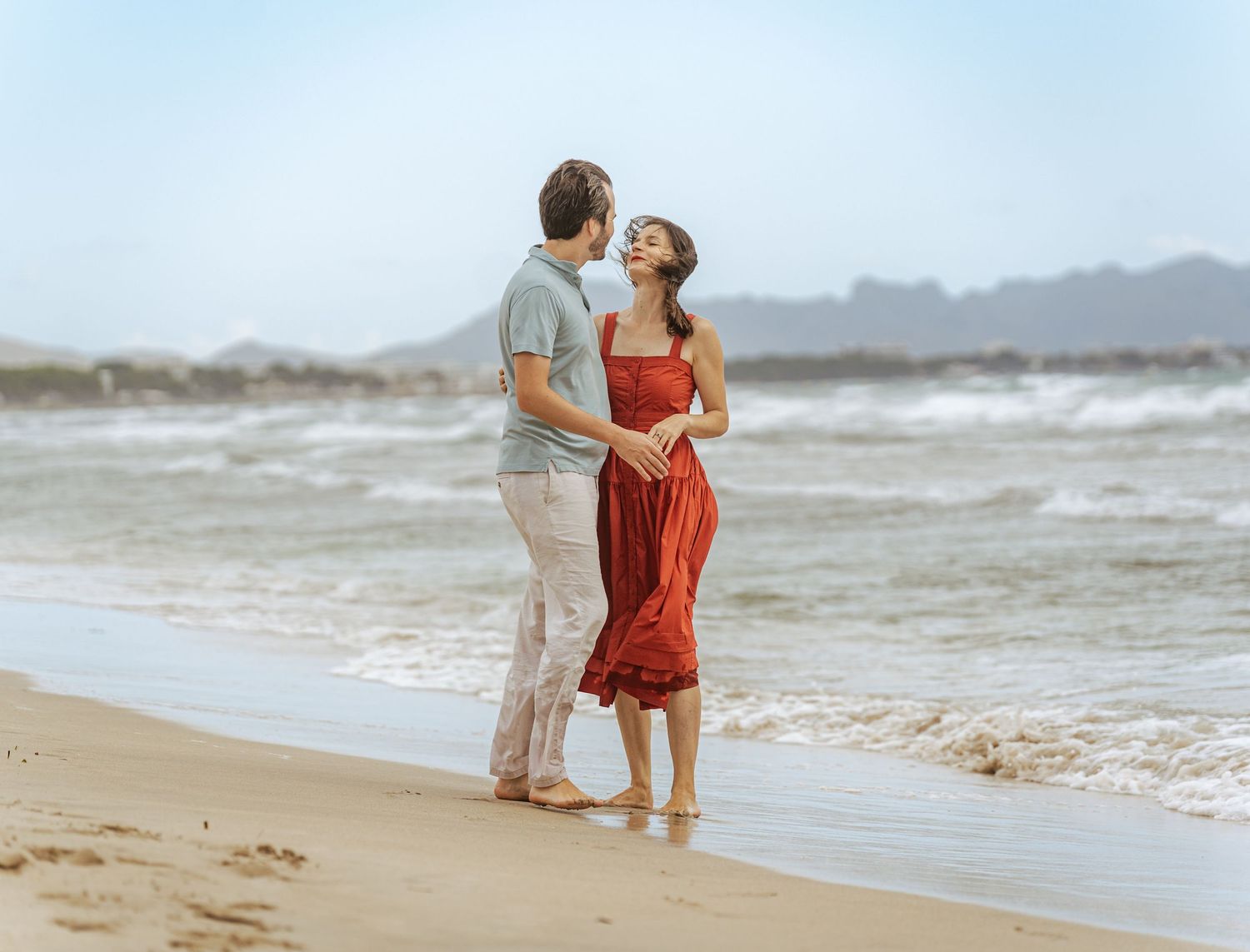 Happy couple holding hands and smiling on a breezy beach, woman in red midi dress, mountains afar.