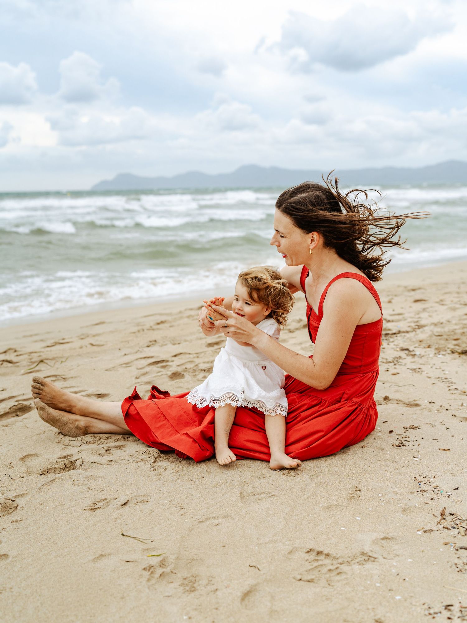 Mother in red dress sitting on beach sand playing with smiling toddler girl in white dress.