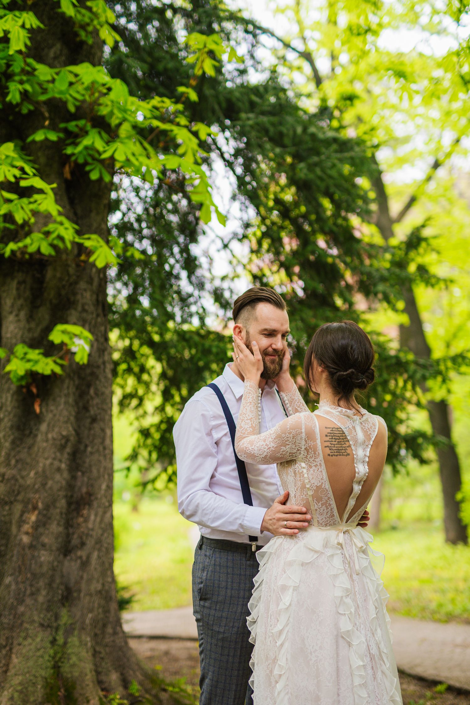 Bride tenderly touches groom's bearded face in a lush green park during outdoor wedding photo session.