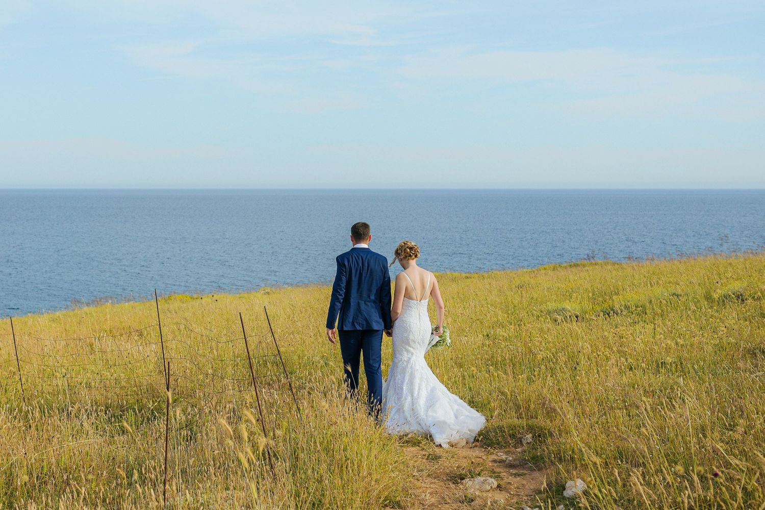 Bride and groom walking together through a golden coastal field overlooking the sea on their wedding day. By Herzenshaus Studio.
