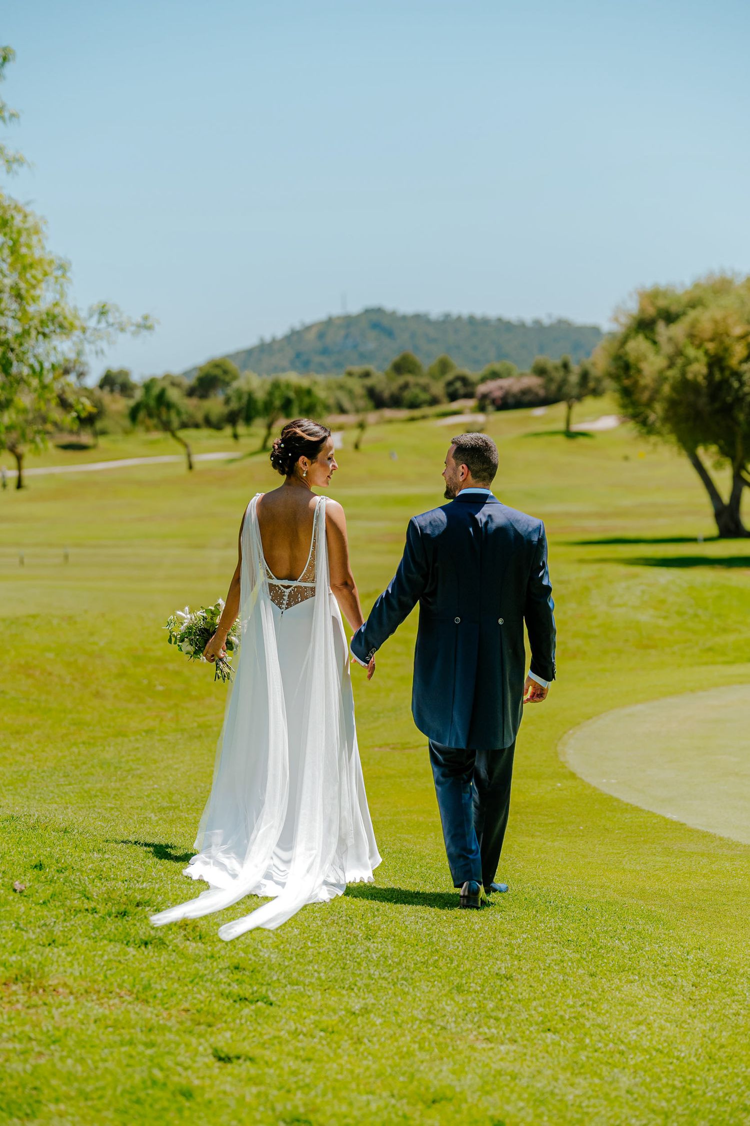 Bride and groom walk hand-in-hand across green golf course with scenic mountain backdrop.