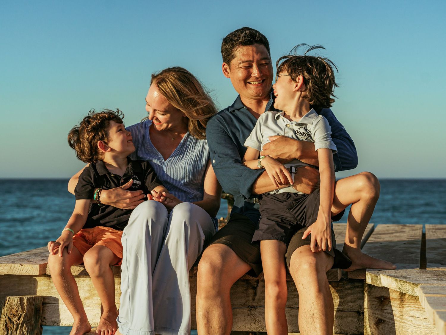 Happy family of four sitting on a pier by the ocean, parents smiling with two young boys at sunset.