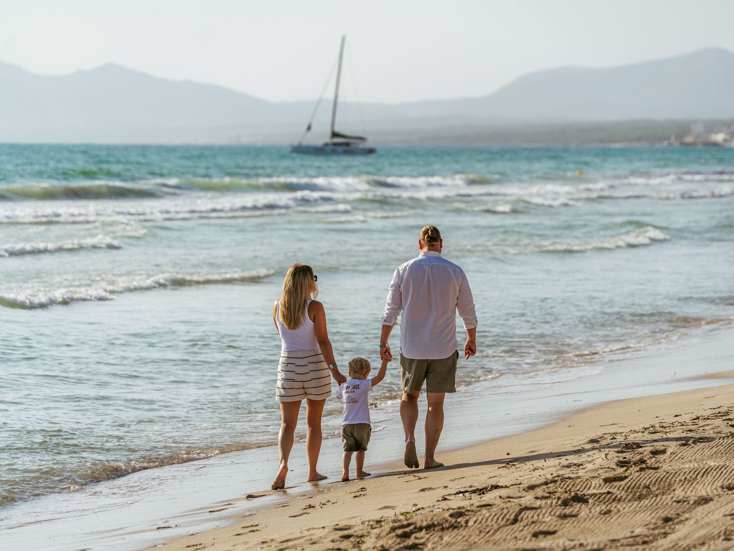Family of three holding hands walking along sandy beach with sailboat and mountains in background.