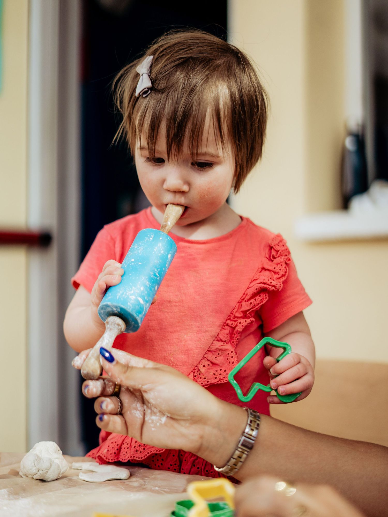 Cute toddler in coral dress chewing on a rolling pin handle while playing with clay dough.