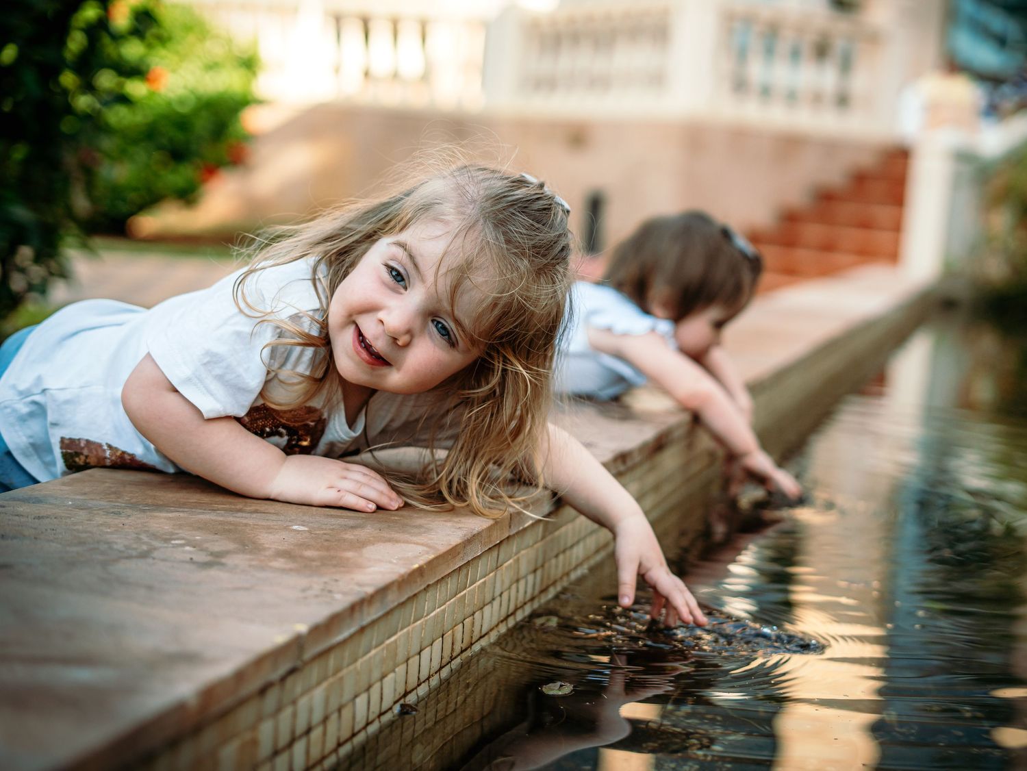Smiling young girl with long hair reaches into a garden water feature while her sibling plays nearby.