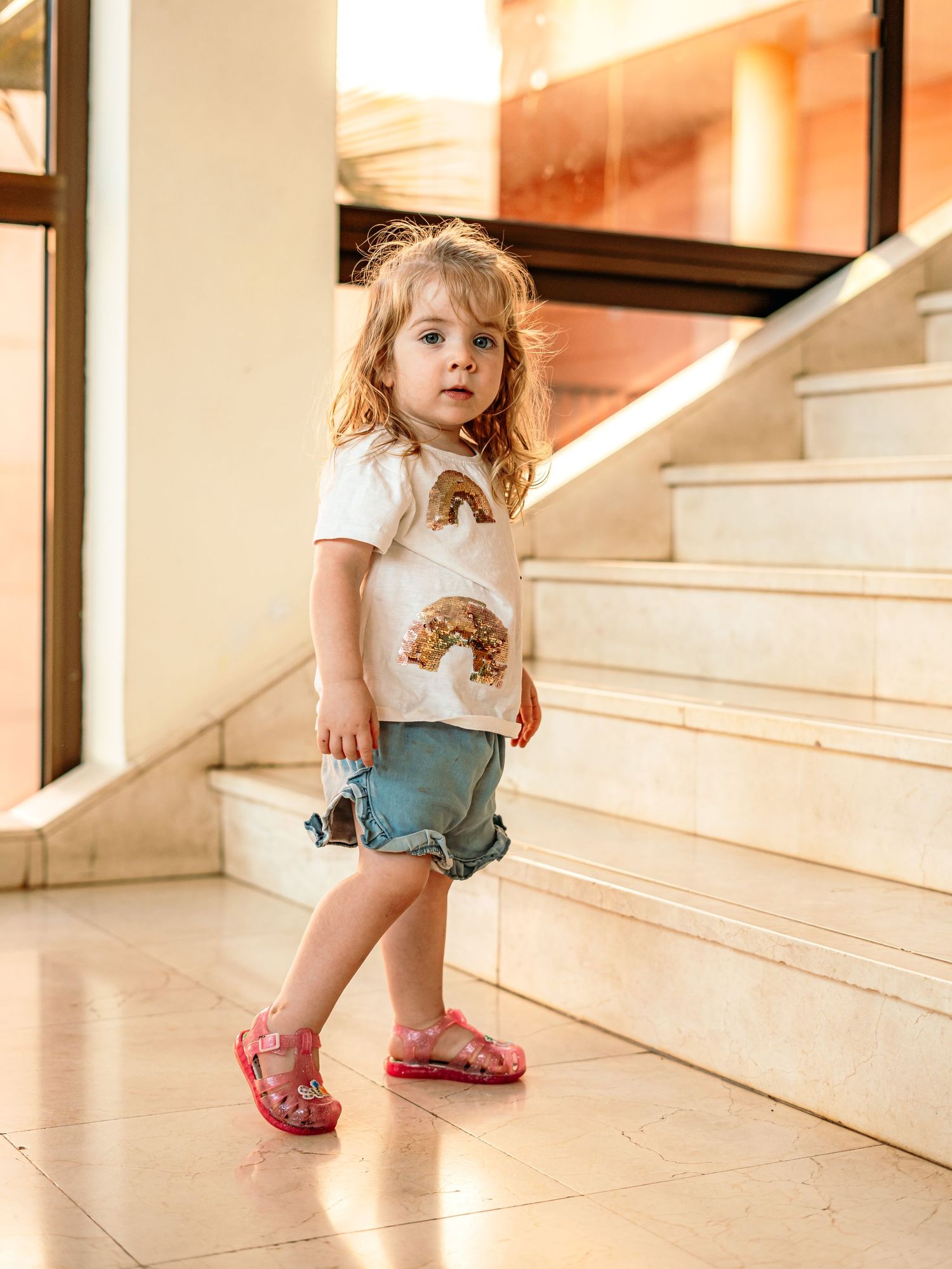 Adorable toddler girl in sequin rainbow t-shirt and denim shorts walking near marble staircase.