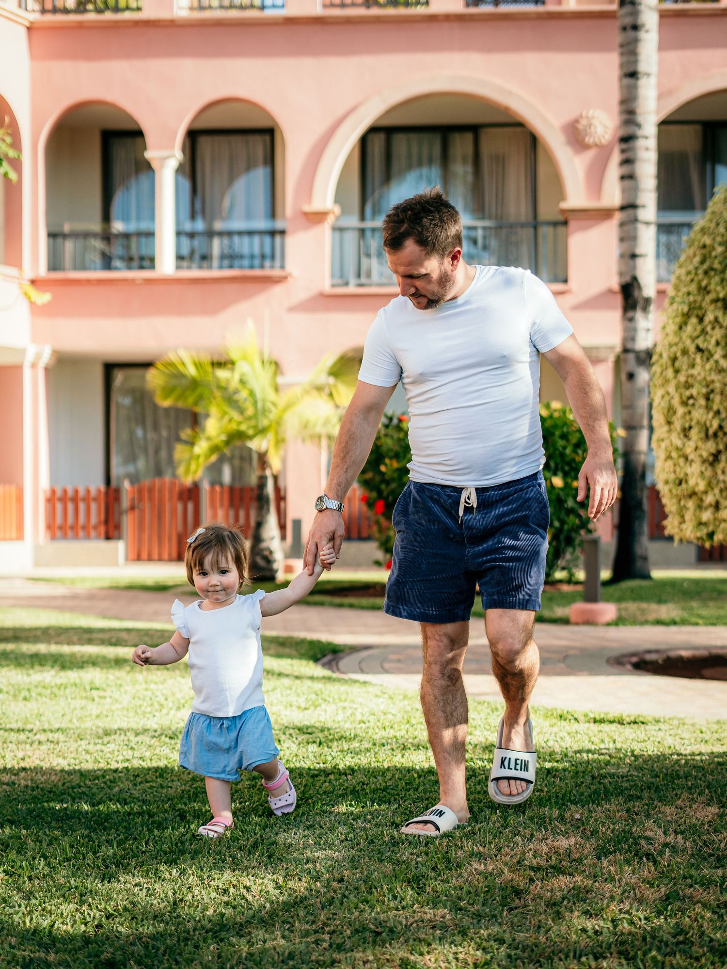 Father in white t-shirt and navy shorts walking hand in hand with toddler daughter on green lawn.